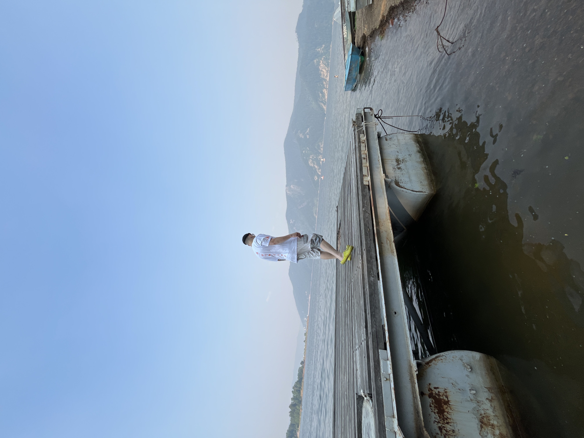 A person wearing a white t-shirt and patterned shorts walks on a wooden pier extending into a calm body of water. The pier is constructed of weathered wooden planks and has a metal railing along its side. In the background, hazy mountains rise from the distant shore. Several small boats are moored near the pier, some are blue, and others are green. The sky is a pale blue, and the overall atmosphere is serene. The scene is likely located in Khmel'niki, Russia, a city known for its natural beauty and water bodies.