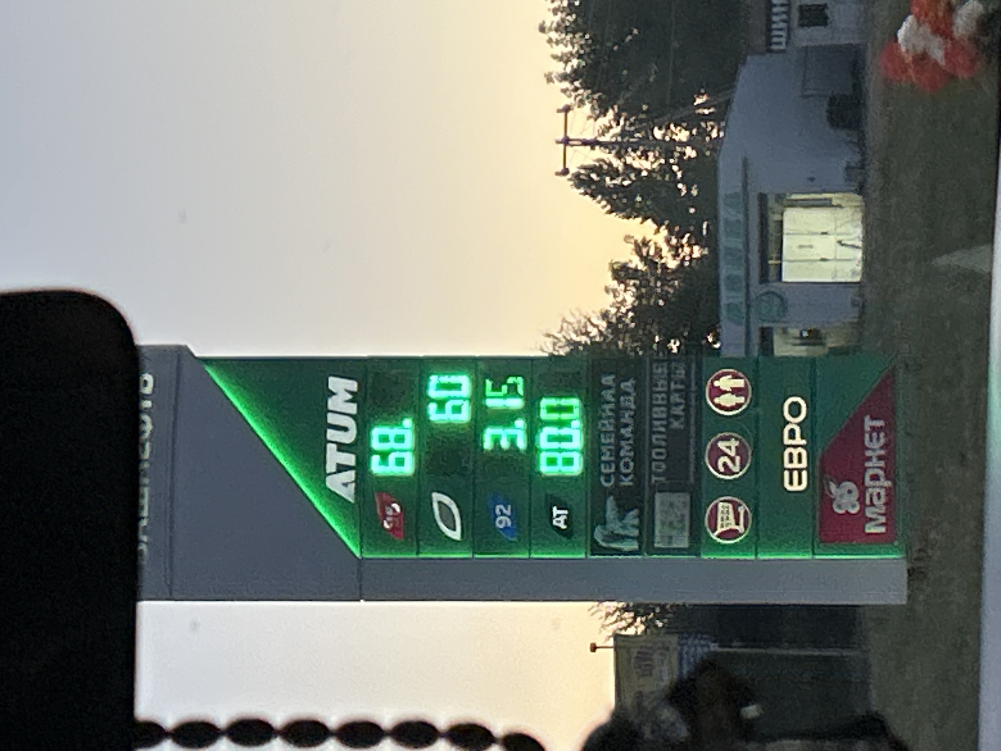 The image captures a gas station price sign in Orsk, Russia. The sign displays prices for different fuel types with illuminated digits. The top section of the sign has the brand name "ATUM" and prices "68." and "60.". Below this, the price "3.15" is shown next to the number "92", indicating the octane rating. Further down, "AT" is displayed alongside "80.0". The sign also includes symbols and text such as "СЕМЕЙНАЯ КОМАНДА" (Family Team), "ТОПЛИВНЫЕ КАРТЫ" (Fuel Cards), and "ЕВРО" (Euro). At the bottom, a red section displays "85" and "Магнет" (Magnet). The gas station appears to be in an outdoor setting with trees and a building with illuminated windows visible in the background. The sky is overcast with a gradient of colors suggesting either early morning or late evening. There are no visible people in the immediate vicinity of the sign, but the overall scene suggests a functioning service station.
