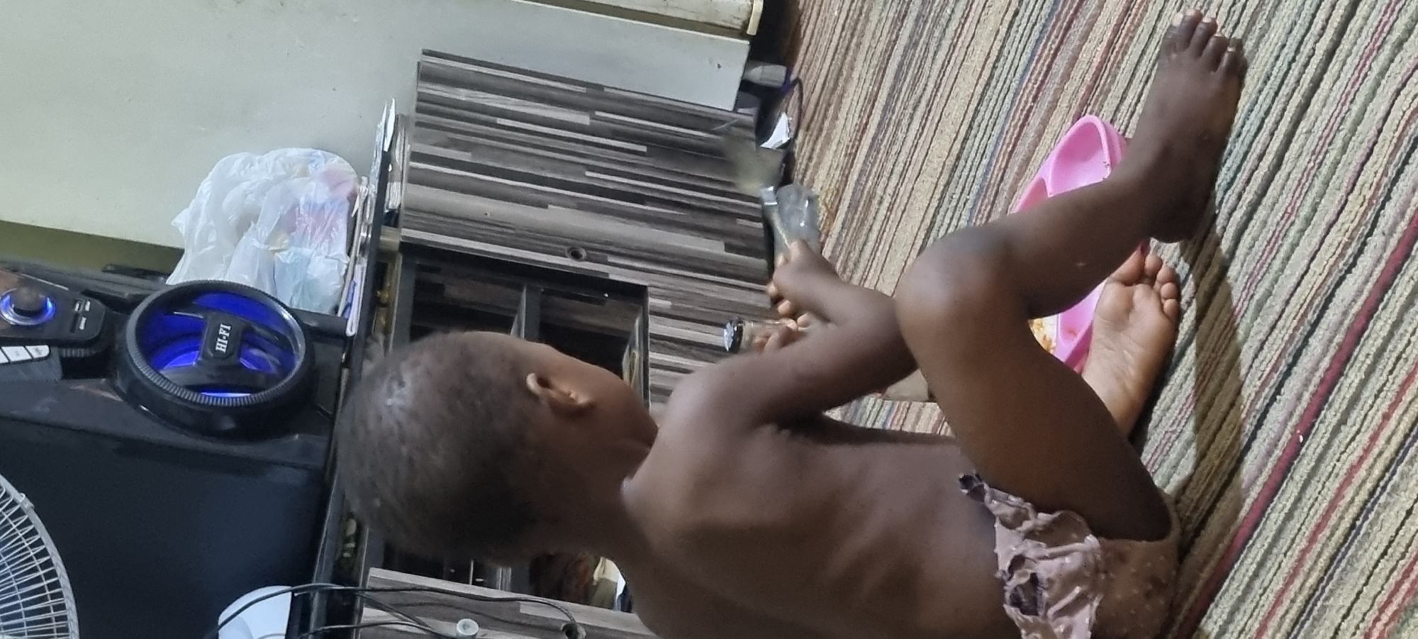 A young child, likely a toddler, is sitting on a striped carpet, facing away from the camera and towards a cabinet. The child is unclothed from the waist up and is wearing patterned shorts. The child is holding a clear plastic bottle and appears to be interacting with it. To the child's right, a pink plastic dish with food is visible. In the background, there is a black speaker system with a blue illuminated fan and the text "HI-FI" on it. Further back is a cabinet with a striped wooden veneer. The environment appears to be indoors, in a residential setting. The lighting suggests it is daytime, possibly in a dimly lit room. The scene is static and lacks any overt signs of distress or unusual activity. Based on the provided context, this image was taken in Lagos, Nigeria.