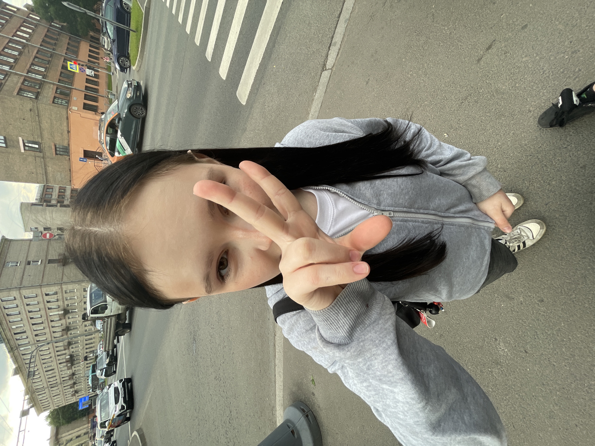 The image is a high-angle selfie, rotated 90 degrees clockwise, depicting a young person on an urban street. The individual has long dark hair and brown eyes, and is wearing a grey hooded zipper jacket over a white t-shirt, dark trousers, and white sneakers with black stripes (Adidas). Their right hand is raised, making a peace sign (V-sign) towards the camera. Part of another person's black shoe is visible on the far right.
The scene is a city street, consistent with Saint Petersburg, Russia. A multi-lane asphalt road with a distinct zebra crossing occupies the foreground and midground. Several vehicles are on the road, including a dark-colored car with an orange, white, and green stripe livery (reminiscent of a taxi service like Yandex.Taxi), a blue sedan, and a white truck further down the street. Multi-story buildings, characterized by brick and stucco facades, line both sides of the street, reflecting typical European urban architecture.
Visible road signs include a yellow diamond-shaped pedestrian crossing sign featuring a red walking figure, and a red circular "No Entry" sign with a horizontal white bar. The lighting indicates it is daytime, likely under overcast conditions as there are no harsh shadows. A keyring with multiple keychains is attached to the person's left side. The image captures a casual street moment, possibly during a walk or sightseeing.