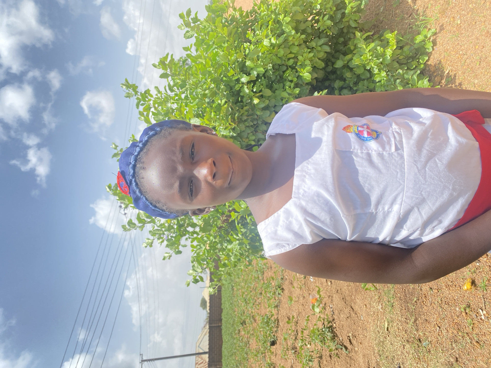 The image depicts a young girl in Turu, Nigeria, standing outdoors on a sunny day. She is wearing a white top with a patch on the chest, a red skirt, and a blue headscarf. Behind her is a green bush and a field of sparse vegetation. Power lines are visible in the background. The ground is a mixture of brown soil and grass. The sky is blue with scattered white clouds.