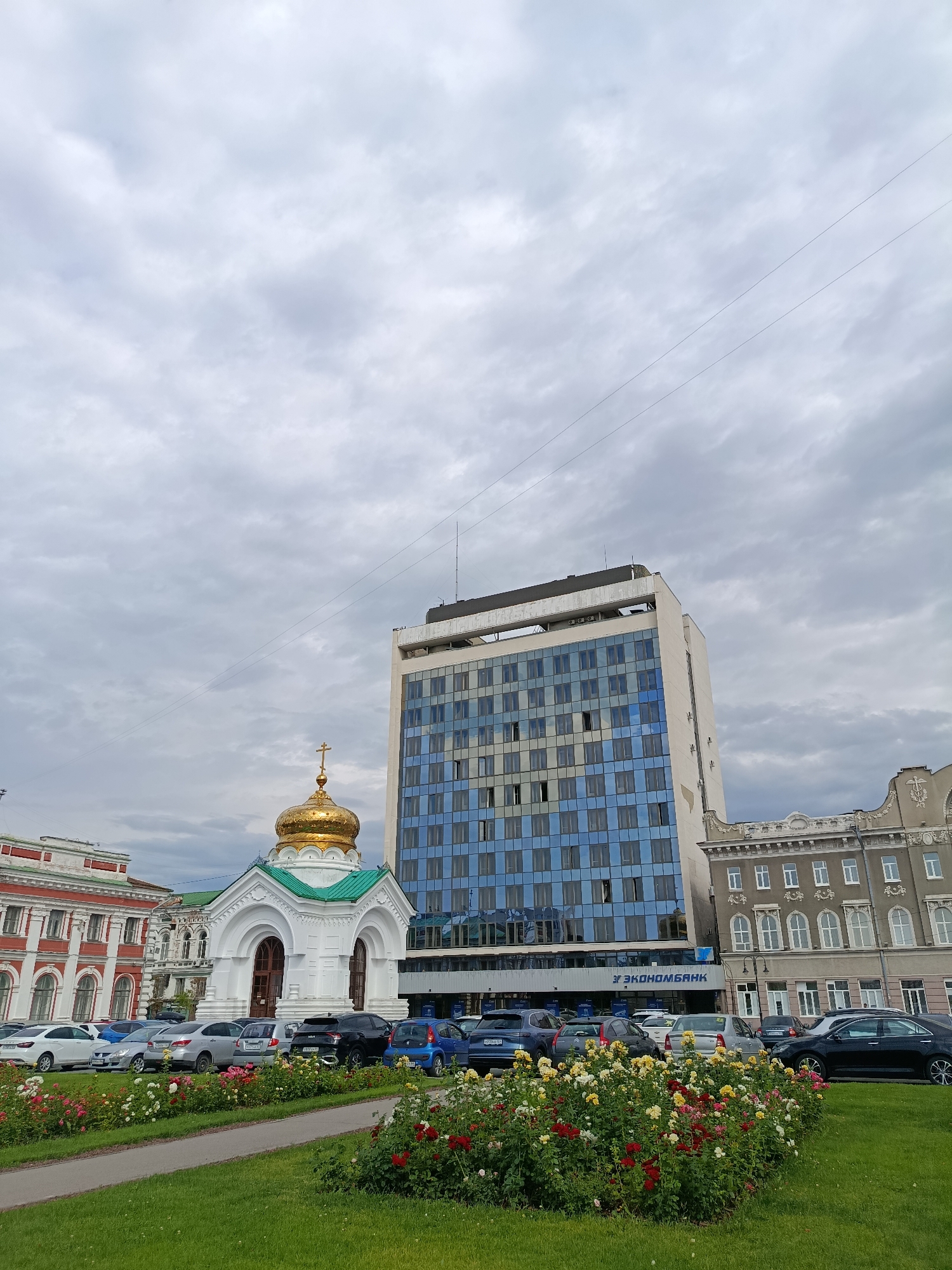 The image captures a street view in Saratov, Russia, featuring a distinctive architectural ensemble. In the foreground, a neatly manicured lawn is bordered by a vibrant flowerbed, predominantly showcasing roses in hues of red, yellow, and white. A paved pathway runs alongside the lawn. Beyond this, a row of parked cars lines the street, including a black sedan, a blue hatchback, several silver sedans, and a white car.
Dominating the mid-ground is a small, white church with a bright golden dome, topped by a cross. The church exhibits traditional Orthodox architectural elements, with arched windows and a green roof. Adjacent to the church stands a tall, modern building with a facade of alternating blue and grey glass panels. The building's upper floors also feature some white structural elements. Below the glass facade, a sign reads "ЭКОНОМБАНК" (Econombank) in blue Cyrillic letters.
To the left of the church, a historic building with red and white detailing is visible, characterized by arched windows. To the right of the modern bank building, another older, grey building with ornate architectural details and numerous windows stands. The sky above is overcast with heavy, grey clouds. The scene depicts a blend of historical and contemporary architecture within an urban setting, likely a public square or street in Saratov.