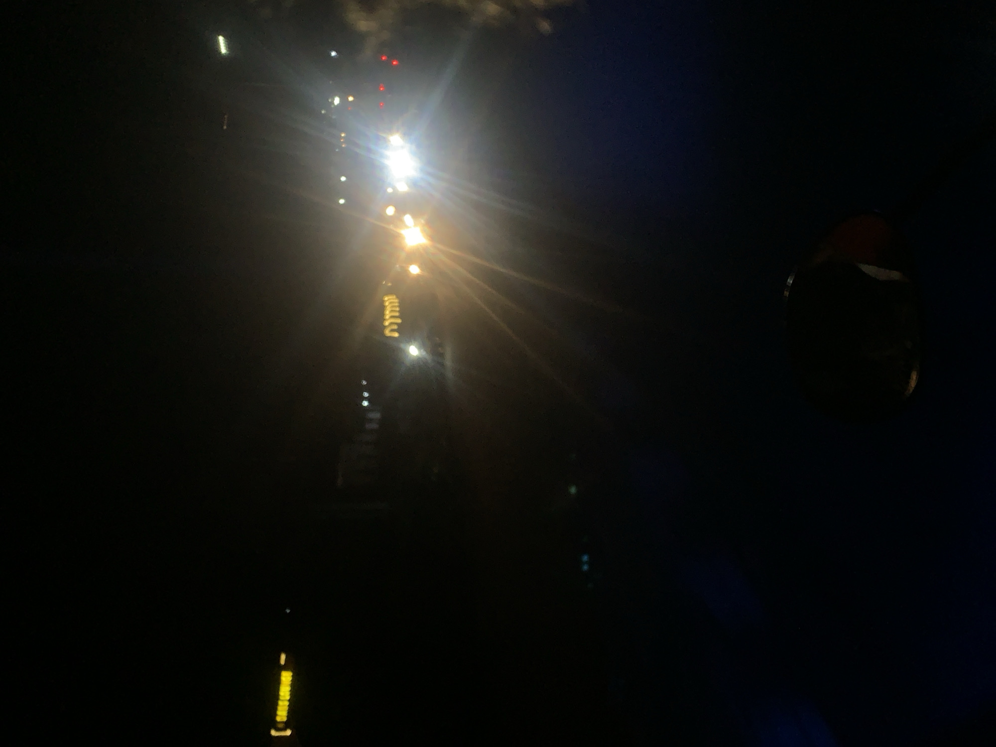 The image shows a dark, nighttime scene in Adankolo, Nigeria. A line of bright streetlights, some with a noticeable glare, stretches into the distance. The lights appear to illuminate a road or walkway. At the bottom left of the image, there are additional, less intense light sources. In the right part of the image, there is a large dark object which is hard to identify clearly.