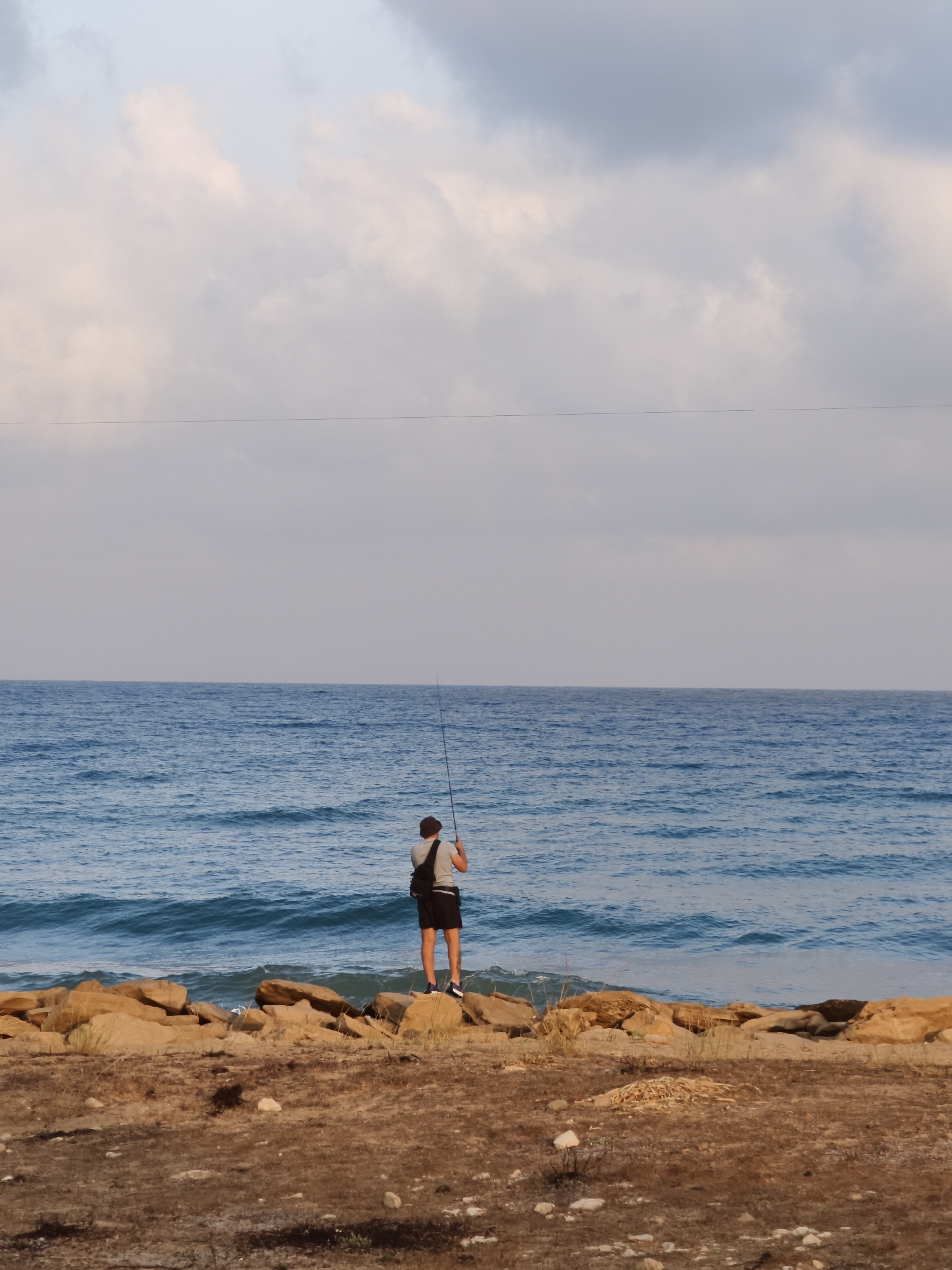 A person stands on a rocky shoreline, facing the sea and holding a fishing rod. The person is wearing a dark hat, a light-colored t-shirt, and dark shorts. A dark bag is slung over their shoulder. The sea is a vibrant blue with moderate waves, and the sky above is filled with pale clouds. The foreground consists of a dry, sandy area with scattered stones and dry vegetation. The scene appears to be on the coastline of Nahariyya, Israel.
