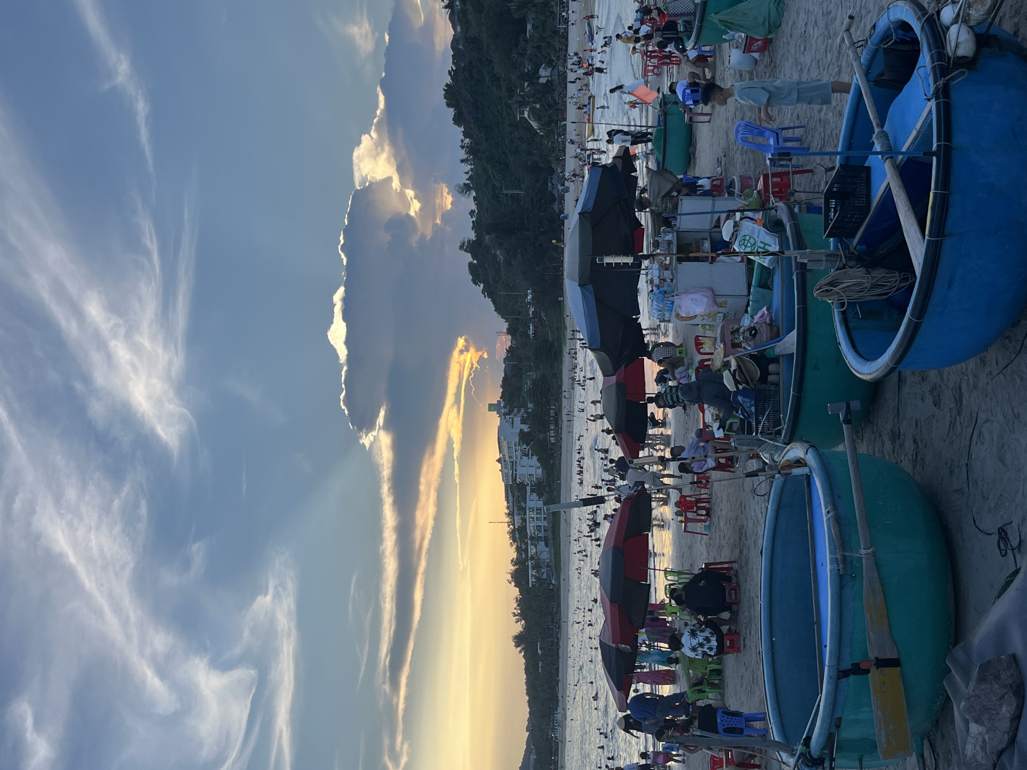 The image depicts a crowded beach scene in Phu Thuy Ward, Vietnam, during what appears to be late afternoon or early evening. The sky is filled with dramatic clouds, with the setting sun casting a warm glow through a break in the clouds, illuminating the water and a portion of the sky with golden and orange hues. The beach is populated with numerous people swimming, sunbathing, and relaxing under beach umbrellas. In the foreground, several round, basket-like boats, common in Vietnamese coastal areas, are pulled up onto the sand. Some people are seated at tables set up on the beach, suggesting food or drink vendors are present. The background features lush green hills with buildings nestled among them, and a hazy shoreline with more people and beach umbrellas. The overall atmosphere is one of a lively and popular tourist destination.