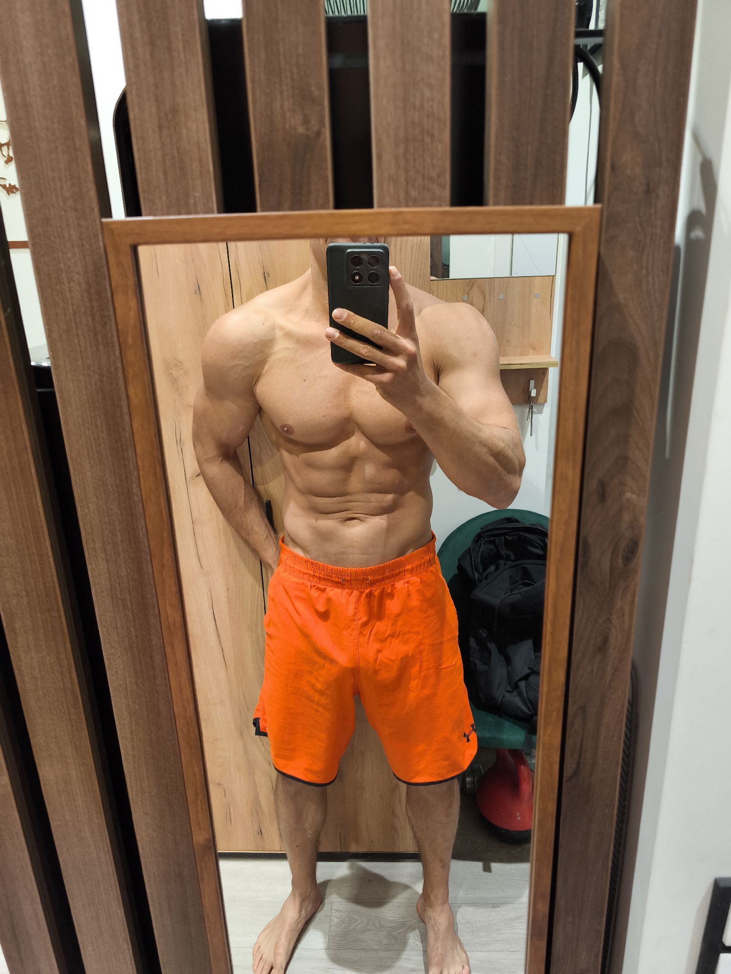The scene is depicted in what is asserted to be a private residence in Moscow, Russia. A shirtless male, approximately 20-35 years old, with a muscular physique and visible abdominal definition, stands barefoot, capturing a mirror selfie. He is wearing bright orange athletic shorts with a thin black trim. His right hand holds a black smartphone, featuring a prominent multi-lens camera array, positioned to photograph his reflection. His left hand rests casually on his left hip. The subject occupies the central portion of a large, rectangular mirror framed with medium brown wood.
The mirror reflects a room interior characterized by warm wood tones. Behind the subject, a light brown wooden door or wall panel is visible, exhibiting a distinct vertical grain pattern. To the right of the subject's reflection, a small, light-colored wooden shelf protrudes from the wall, with two keys hanging from a white hook. Below the shelf, a dark green, upholstered stool or small chair is partially visible, draped with a crumpled black garment, possibly a jacket. Adjacent to the stool on the floor, a red kettlebell is discernible. The floor within the reflection is a light grey or pale wood laminate.
Outside the mirror's frame, the immediate environment includes a wall to the left covered with vertical dark brown wooden slats, creating a striped pattern against a lighter background. To the right of the mirror, a plain white wall is visible. Above the mirror, more of the vertical wooden slats are present. A partial view of a black bicycle frame or wheel is subtly reflected in the upper right corner of the mirror, just outside the primary composition.