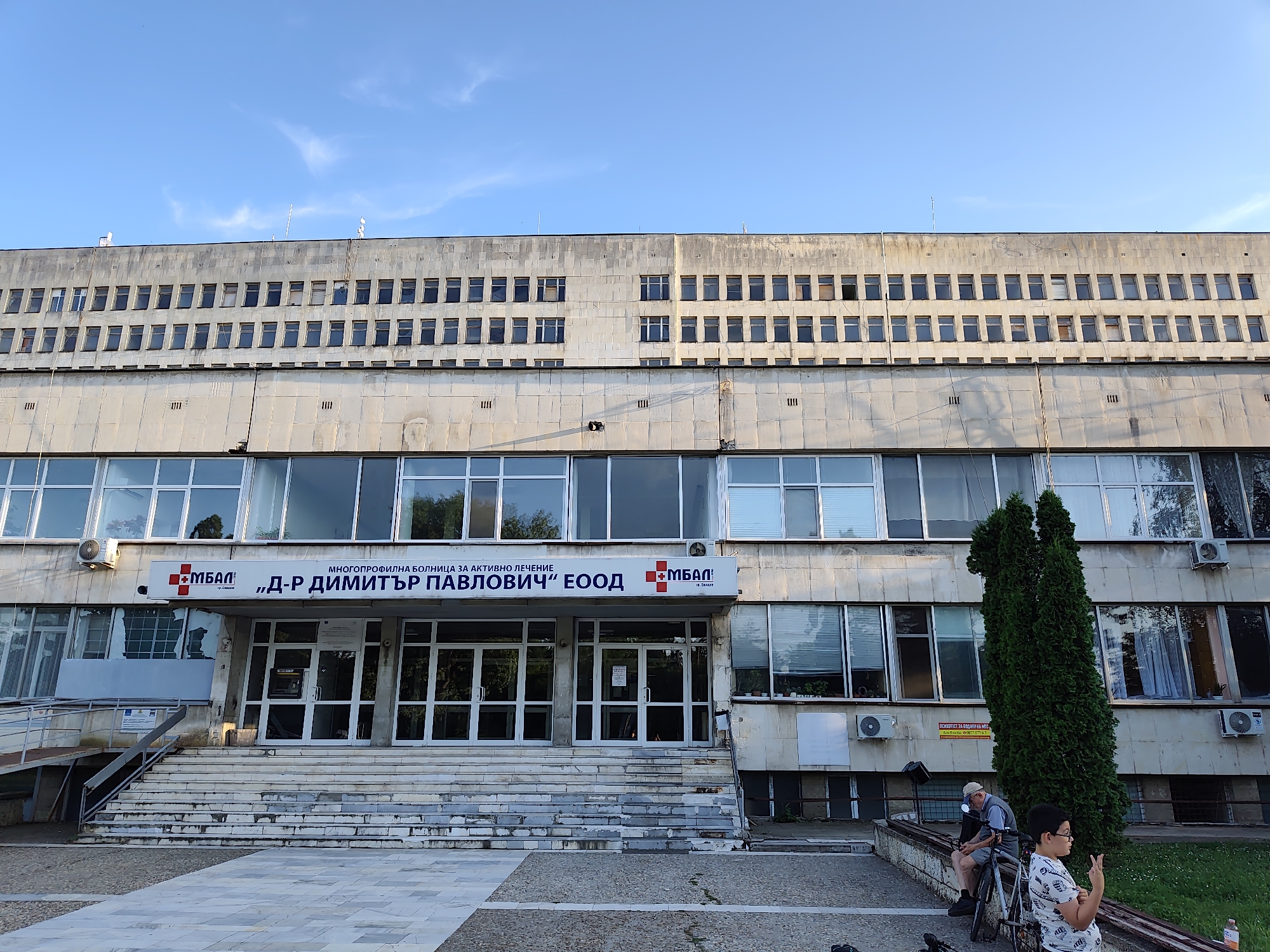 The image shows the exterior of a large, multi-story building, likely a hospital, with a sign that reads "МБАЛ Д-Р ДИМИТЪР ПАВЛОВИЧ" and "МНОГОПРОФИЛНА БОЛНИЦА ЗА АКТИВНО ЛЕЧЕНИЕ" which translates to "Multi-profile Hospital for Active Treatment 'Dr. Dimitar Pavlovich'". The building is constructed of concrete and has numerous windows. The entrance features a wide set of stairs and glass doors. A large evergreen tree stands to the right of the entrance. In the foreground, two people are visible: an older man sitting on a bench next to a bicycle, and a young boy standing and looking towards the camera, holding up two fingers. The scene appears to be set during daylight, with a clear blue sky overhead. The building is located in Svishtov, Bulgaria.