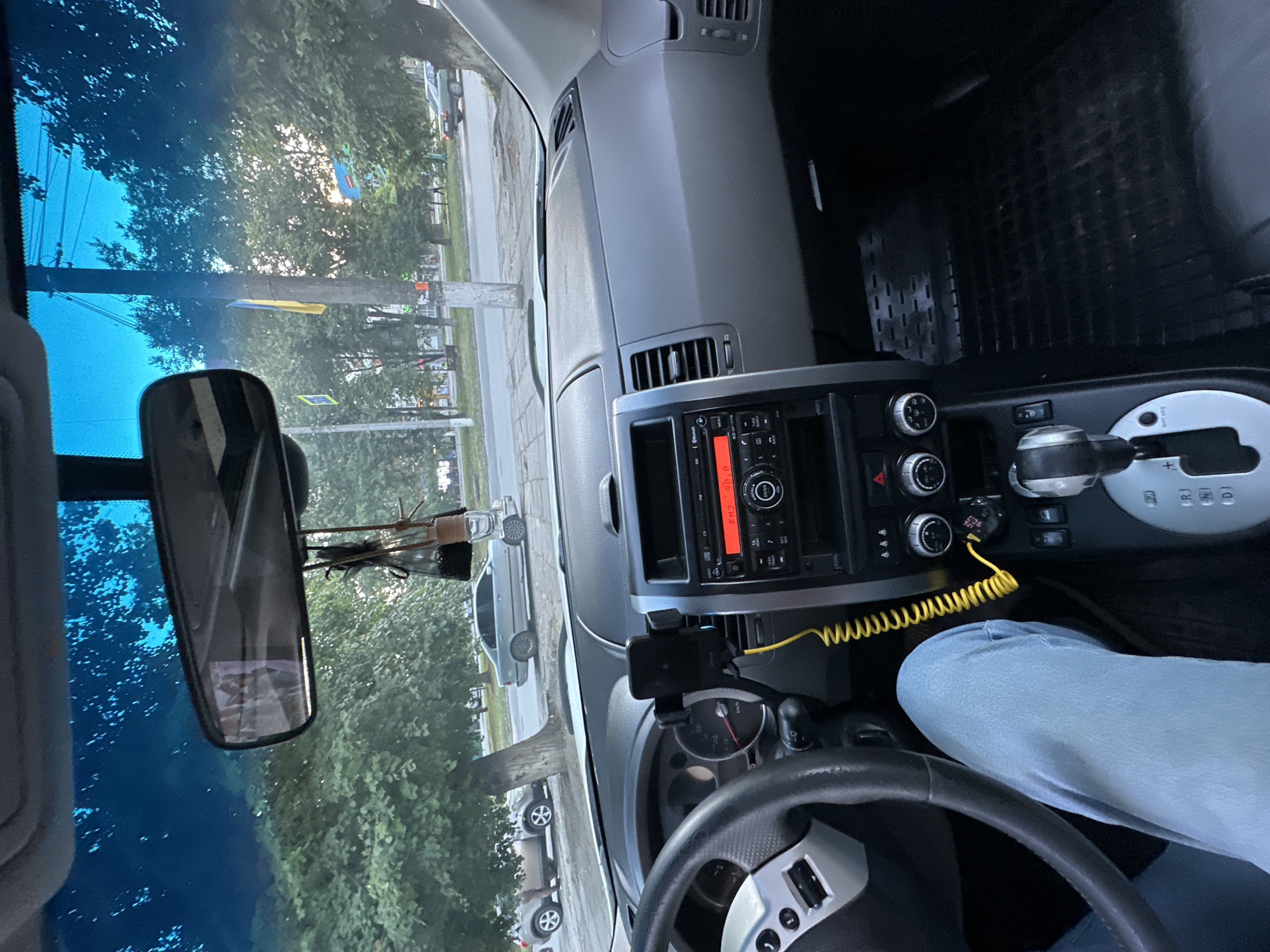 The image is taken from the interior of a car, looking out through the windshield. The dashboard is visible in the foreground, with a car stereo system, climate controls, and a mobile phone mounted on a holder. A person's legs, wearing light blue jeans, are visible on the left side of the frame. The steering wheel is also partially visible. Outside the car, trees and a street with cars are seen. In the background, a yellow and blue flag is hanging from a pole, which is a clear indicator of the location being Ukraine. The Ukrainian flag is also visible on a building in the background. The presence of the Ukrainian flag, combined with the general urban and suburban environment depicted, suggests the photo was taken in Kamianets-Podilskyi, Ukraine. The lighting suggests it is daytime, possibly late afternoon or early evening.