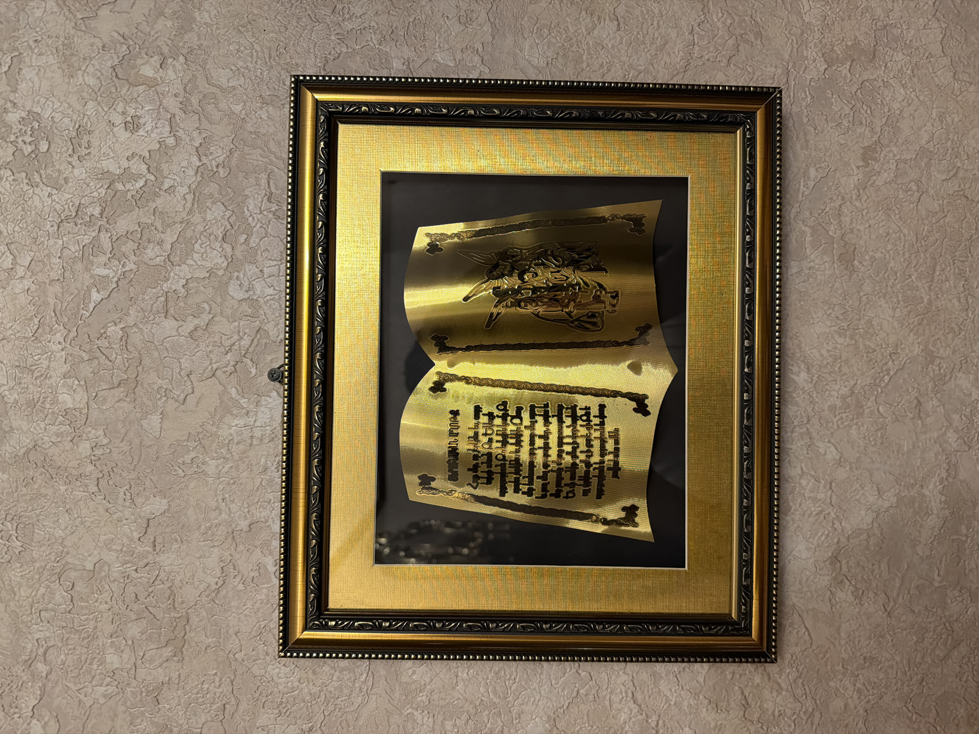 A framed, ornate, rectangular picture hangs on a textured wall. The frame is gold with black decorative elements. Inside the frame is a piece of gold-colored metal shaped like a scroll. Etched onto the metal scroll are designs and text. The lighting in the scene suggests an indoor environment. The wall has a neutral color and a rough, textured surface. The image provides no definitive information to confirm its location in Krasnoyarsk, Russia.