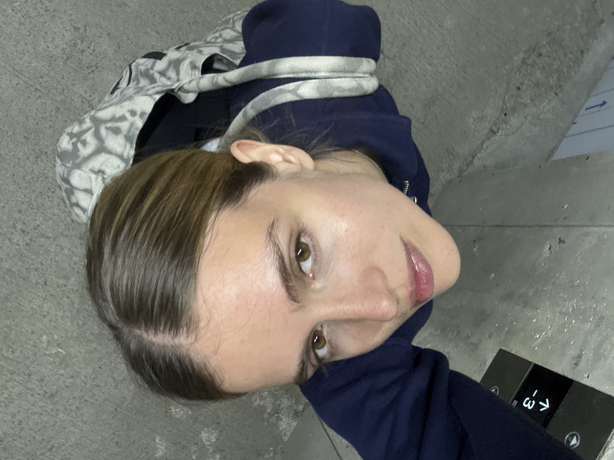 A high-angle shot shows a young woman looking up at the camera. She has light brown hair and brown eyes. She wears a dark blue hoodie, and part of a patterned bag is visible near her head. The woman appears to be in an elevator, as evidenced by an elevator control panel located in the lower right corner of the frame. The background consists of the interior wall and floor of the elevator, which are a neutral concrete color. The location appears to be Unknown, Unknown.