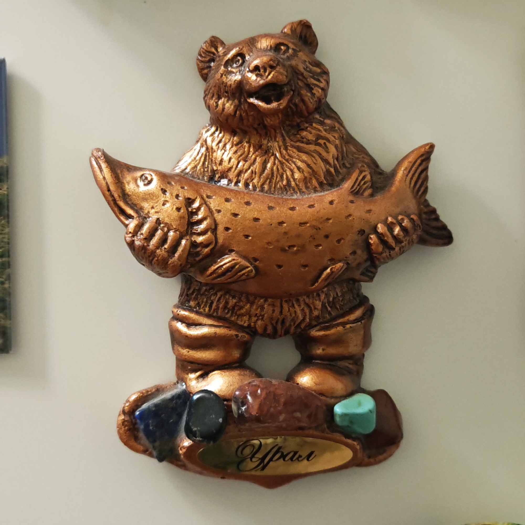 This image captures a close-up of a metallic-looking souvenir or magnet, likely from Novo-Nikol'skoe, Russia. The central subject is a bear figure, cast in a bronze or copper tone, standing upright and holding a large fish horizontally across its body with both paws. The bear has an open mouth, giving it an expression of joy or perhaps a roar, and features detailed fur texture. The fish also displays textured scales or spots.
The base of the figurine is decorated with several small, polished stones of various colors, including deep blue, black, reddish-brown, and turquoise, suggesting mineral wealth. Embedded in the base is a gold-colored oval plaque with the word "Урал" (Ural) written in elegant Cyrillic script, indicating the region this souvenir represents. The object is presented against a plain, light-colored background, characteristic of an indoor setting, possibly on a refrigerator or wall. The overall appearance suggests a tribute to the wildlife and natural resources of the Ural Mountains.