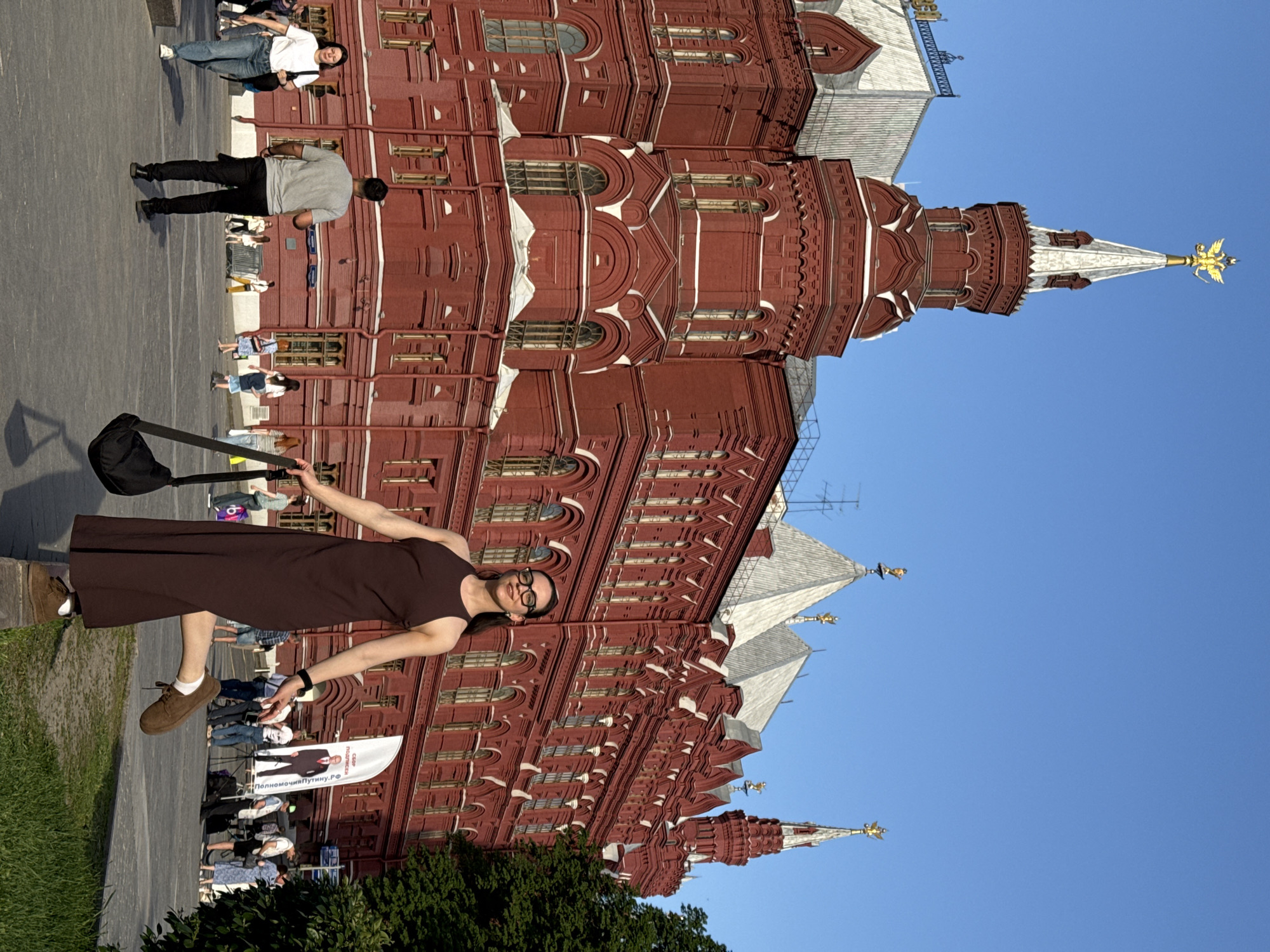 A woman in a brown dress and glasses stands with arms outstretched in front of the State Historical Museum in Moscow, Russia. She is holding a black bag. Several other people are nearby, including two lying on the ground. A white banner with text and an image of a man in a suit is visible near the bottom of the frame. The sky is blue.