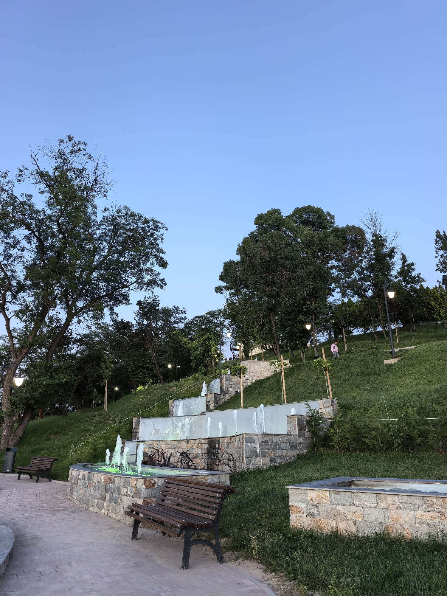 The image depicts a terraced park area with a cascading fountain. The fountain features multiple levels with water jets, some illuminated with green light. Stone walls and pathways define the various levels. Two wooden park benches are visible on the paved area in the foreground. Lush green grass covers the sloping terrain. Several trees of varying sizes are scattered throughout the scene, with a prominent large tree on the left. The sky is clear and blue, suggesting it is daytime. The park appears to be located in Svishtov, Bulgaria, based on the provided context.