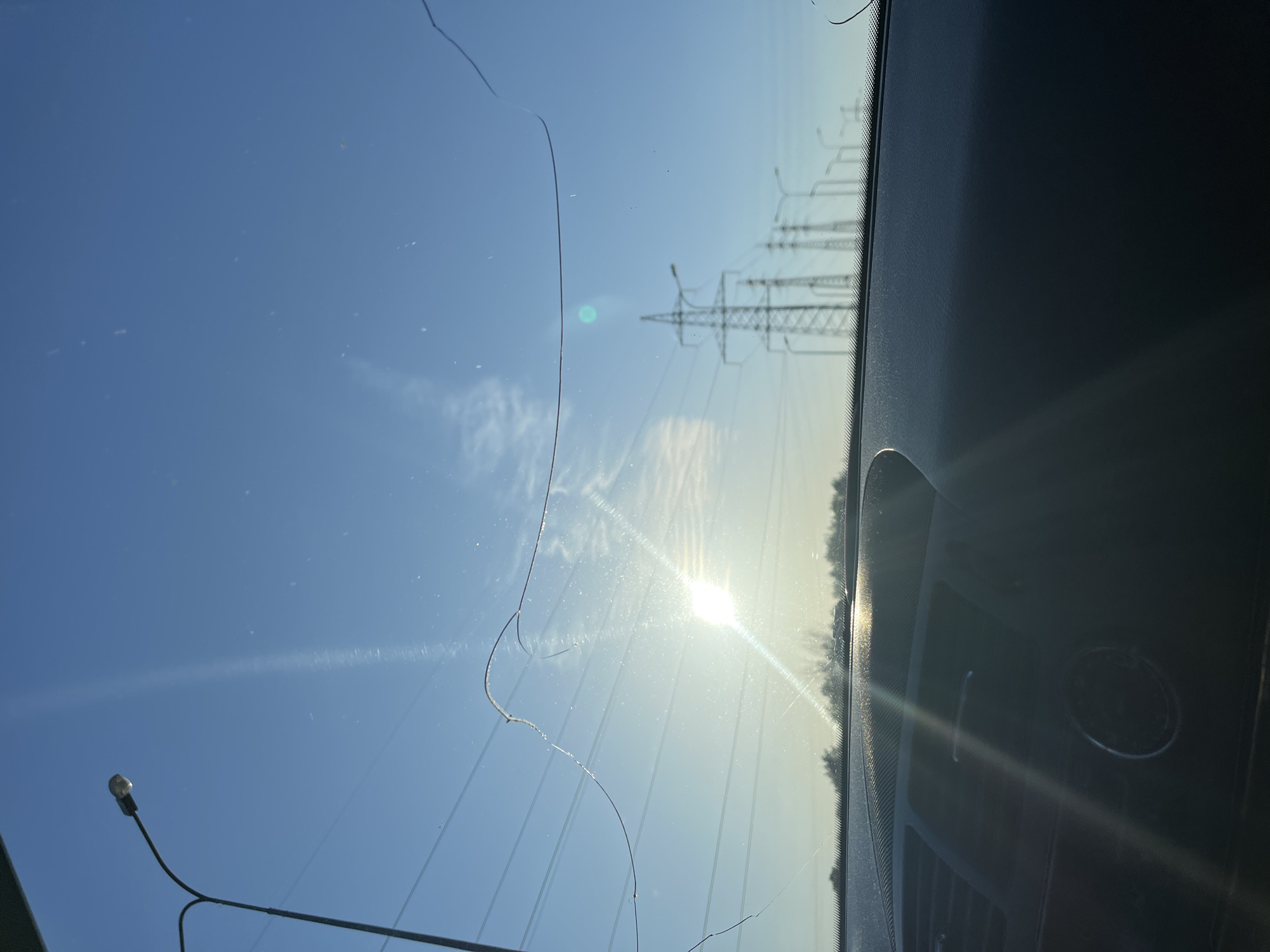The image shows a cracked windshield from inside a vehicle, with a view of a clear, blue sky and the sun. Visible through the windshield are power lines and supporting towers, as well as a street lamp. The right side of the image shows the vehicle's dashboard, and the sun is reflecting off the glass. There is no indication of this being Chaikovsky, Russia.