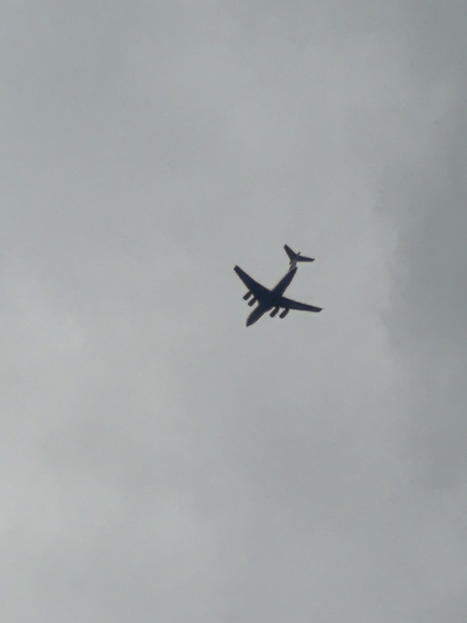 A silhouette of a large airplane is visible against a cloudy, overcast sky. The plane is depicted from below, with its wings extended and tail fin prominent. The aircraft appears to be a four-engine jet, possibly a cargo plane or a passenger jet, though specific model details are not discernible due to the silhouette and image quality. The sky is a uniform shade of grey, suggesting it is daytime with significant cloud cover. No other objects or elements are present in the frame. The image does not contain any information that would allow for the identification of a specific location within the city of Verkhnyaya Pyshma, Russia.
