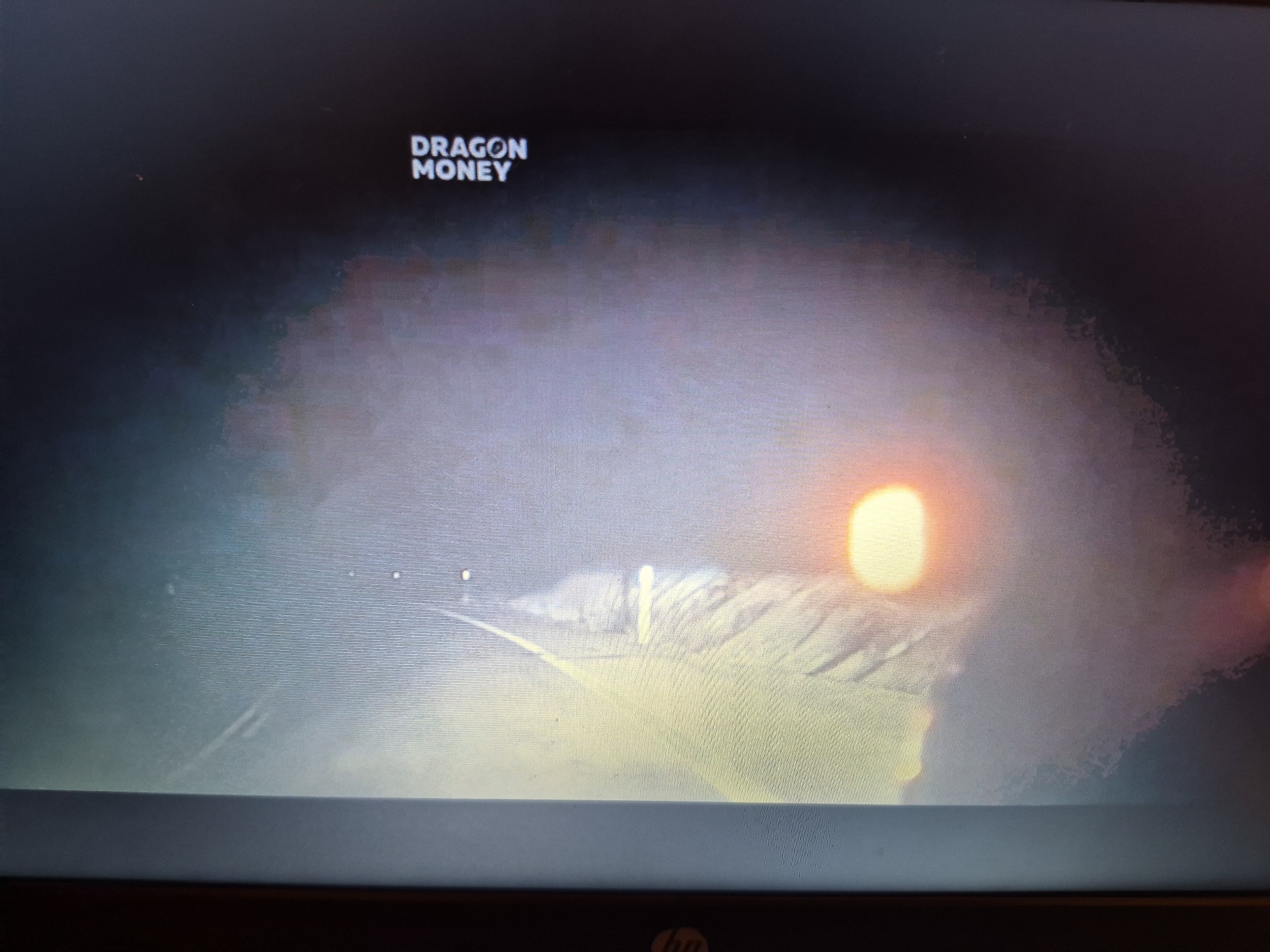 The image displays a computer screen showing a title card for "DRAGON MONEY" overlaid on a sunset scene. The sunset features a bright, circular sun with a halo effect, casting a golden glow over what appears to be clouds or mist. The overall lighting is dim, with a gradient from dark at the top to lighter hues towards the bottom where the sun is situated. The screen itself appears to be a laptop display, with a portion of the keyboard bezel visible at the bottom of the frame, including an HP logo. There are no identifiable people or specific landmarks of Sandwell visible.