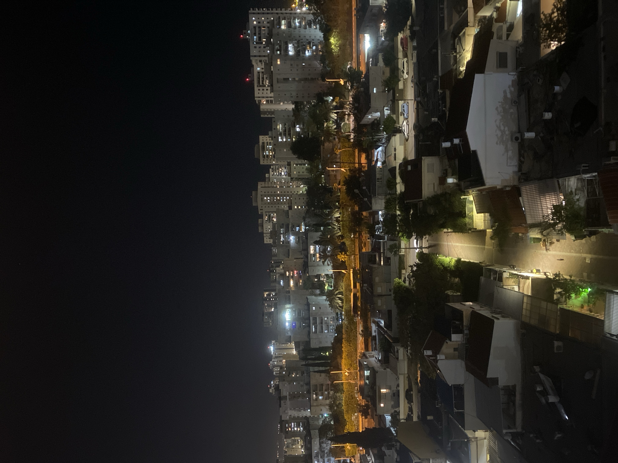 The image shows an aerial night view of Kiryat Bialik, Israel. Tall apartment buildings are visible on the left, transitioning to lower residential structures and streets on the right. Streetlights illuminate the streets and buildings. No people are directly visible, but the presence of lit windows suggests occupancy. Cars are parked along the streets. The sky to the left is predominantly dark.