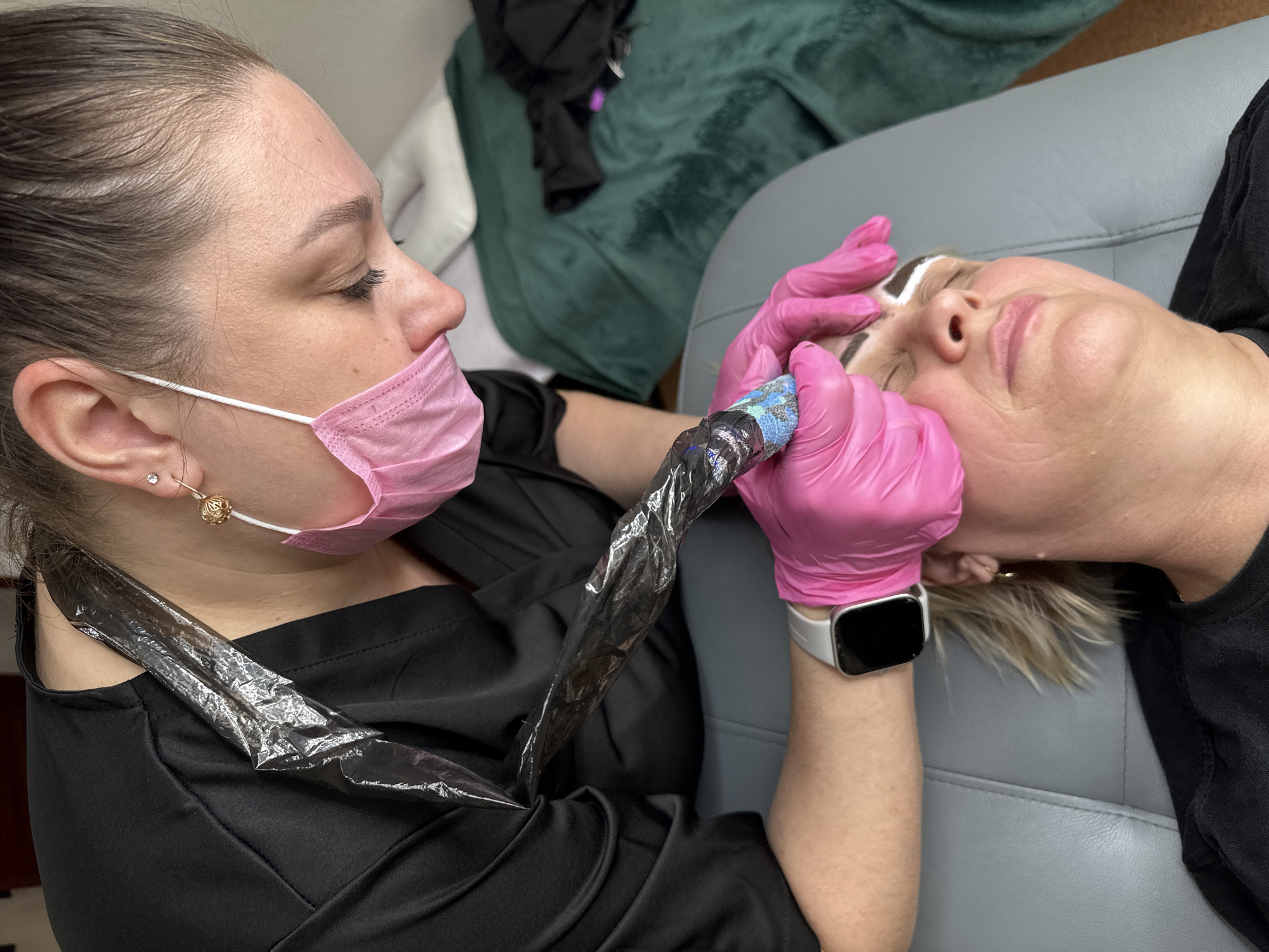 A medium indoor shot depicts a woman with dark hair in a ponytail and a pink face mask performing a cosmetic procedure on another woman lying on a grey upholstered surface. The woman performing the procedure wears pink gloves and is holding a tattoo machine with a cord covered in clear plastic wrap. The machine is poised near the brow area of the woman lying down. The second woman's face is prepped with white markings around the brow area. The woman performing the procedure appears to be looking at the recipient of the service. The woman receiving the service has her eyes closed. A black garment and green cushion are visible in the background. A smartwatch is visible on the wrist of the woman giving the service. There is no contextual information indicating this scene occurred in Chaikovsky, Russia.