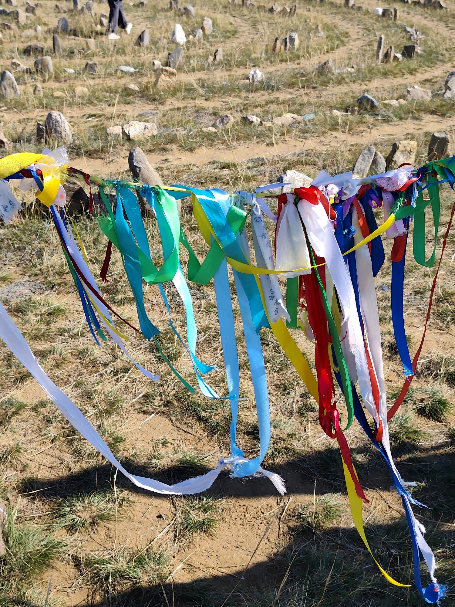 The image depicts a cemetery in Kharantsy, Russia, where colorful ribbons are tied to a horizontal wire, possibly as offerings or prayers. The cemetery appears to be on a grassy hillside dotted with tombstones, some of which are weathered stones. The ribbons are of various colors, including white, blue, green, yellow, and red, and are fluttering in the wind. In the background, a person's legs are visible, walking through the cemetery. The ground is dry and covered with sparse grass and dirt. The scene suggests a solemn yet vibrant atmosphere, with the ribbons adding a touch of color to the otherwise natural and somewhat stark environment.