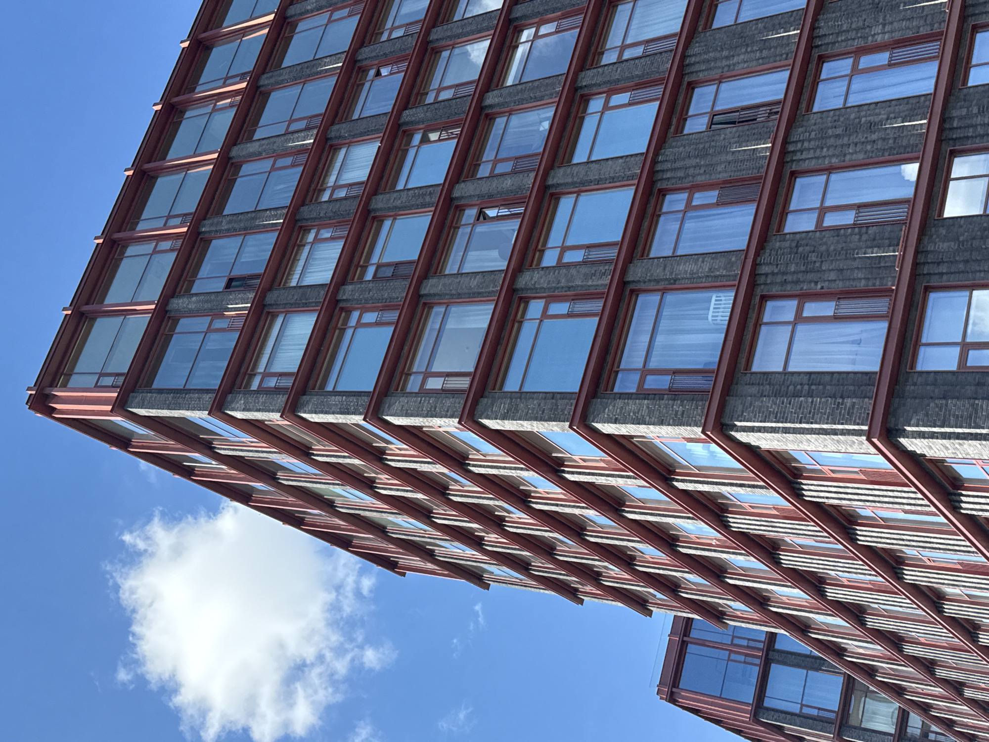 An exterior, low-angle, full shot captures the corner of a modern, multi-story building with a dark gray brick facade and red metal trim around its numerous large windows. The building is viewed from below, looking upwards towards a bright blue sky with scattered white clouds. The architecture features repeating rows of windows on each level, with some windows featuring small Juliet balconies. To the left and further back, another section of the building, also with red trim and large glass panels, is visible. The scene appears to be an urban environment, likely a residential or commercial building. The image was taken at the Karetny Plaza residential complex in Moscow, Russia.