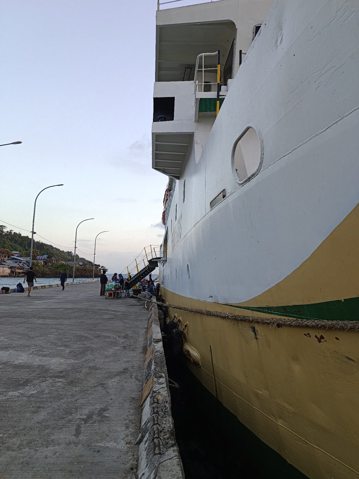 In Mansalean, Indonesia, a large white and yellow ship is docked at a concrete pier. The ship's hull is painted white with a distinctive yellow stripe near the bottom, and a green band runs just above the waterline. The pier is wide and extends into the water, with several streetlights lining its edge.
On the pier, a few individuals are seen. Some are standing near the ship, possibly waiting to board or disembark. Others are walking along the pier, carrying luggage. In the background, a small village with houses is nestled on a green, hilly landscape that meets the water's edge. The sky is overcast, suggesting it might be early morning or late afternoon. The scene appears to be a typical port activity, with people interacting with the docked vessel.