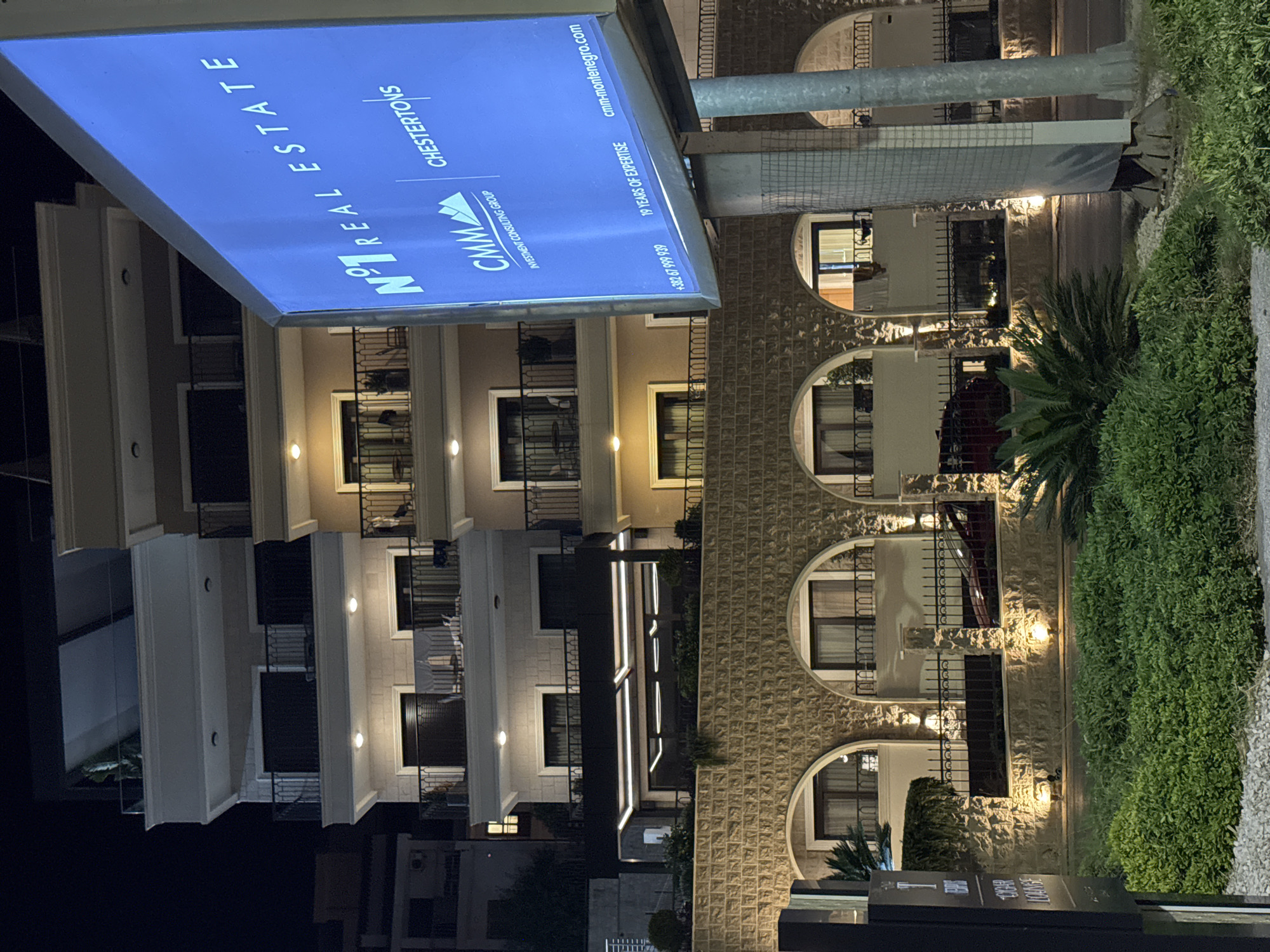 A night-time, low-angle shot captures the facade of a modern building with multiple balconies and arches, illuminated by warm outdoor lighting. In the foreground, lush green bushes and manicured plants are visible, along with a dark sign on the left with some illegible text. Dominating the right side of the frame is a large, illuminated blue billboard displaying advertising text for "N°1 REAL ESTATE" and "CHESTERTONS". The building features a stone facade with arched entrances on the lower level and lighter-colored walls with numerous windows and balconies on the upper floors. The overall impression is one of a well-lit, upscale commercial and residential property in a developed area.