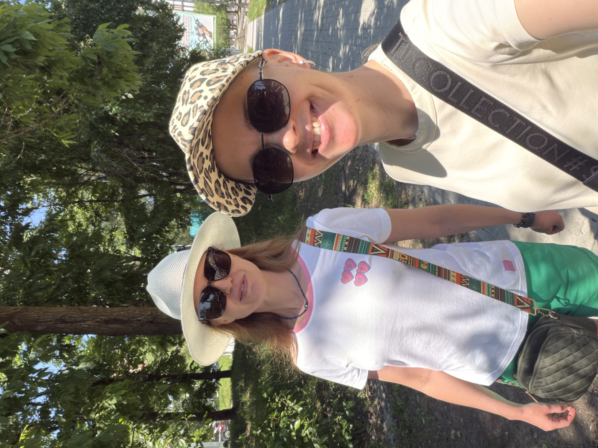 Two women are standing outdoors on a sunny day, possibly in Khabarovsk, Russia. The woman on the left is wearing a white hat, sunglasses, and a white t-shirt with pink heart graphics and the letters "TR" and "MT". She has a colorful patterned strap across her body and is carrying a quilted green bag. The woman on the right is wearing a leopard-print cap, sunglasses, and a light-colored t-shirt with a black strap across her body. She is smiling and wearing hoop earrings. They are standing on a sidewalk with trees and greenery in the background.