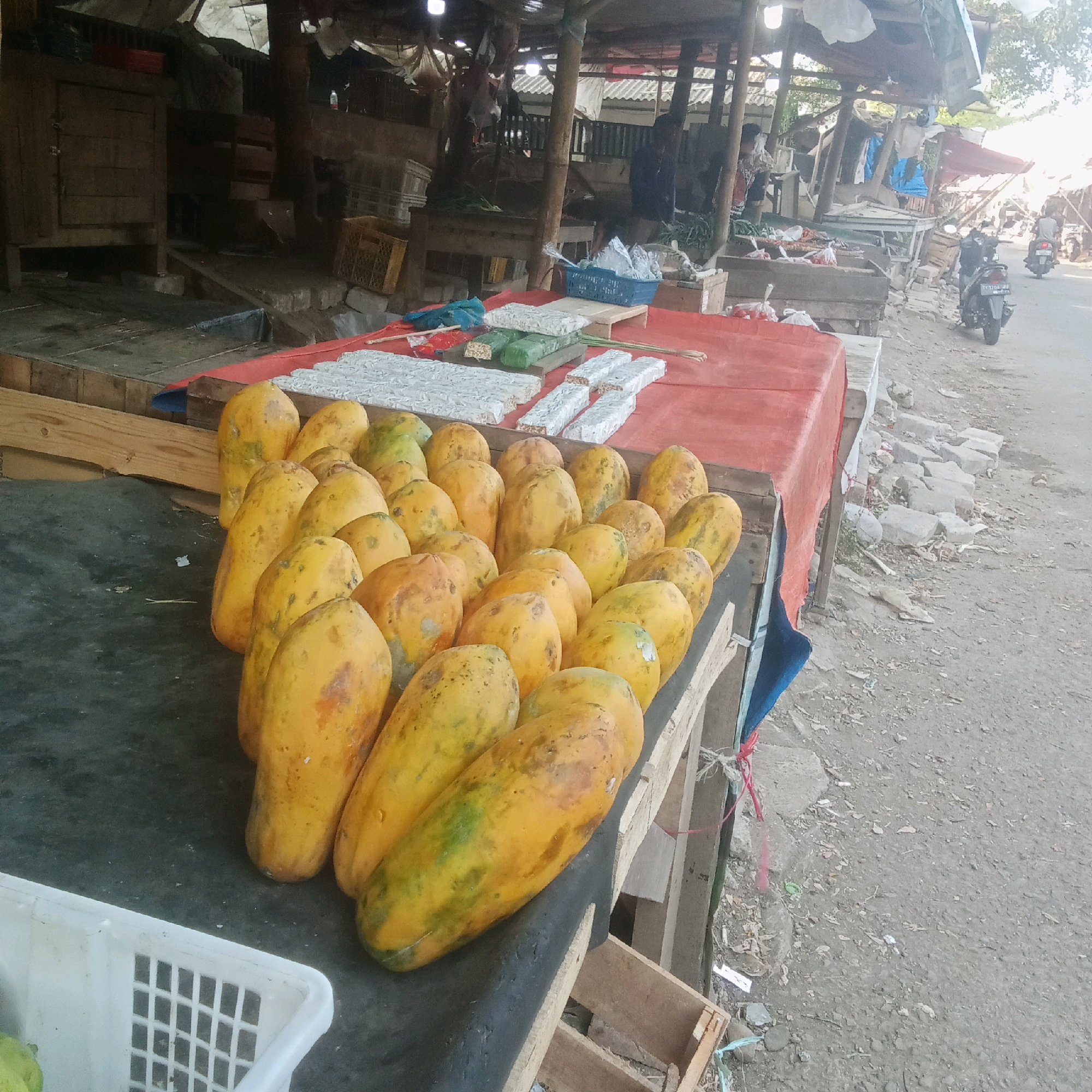 The image depicts an outdoor market scene in Karawang, Indonesia. In the foreground, a collection of ripe papayas, predominantly yellow with some green streaks and brown blemishes, are displayed on a dark surface, likely a stall. Behind the papayas, several rows of white, rectangular items, possibly processed foods like tempeh or dried fish, are arranged on a red tablecloth. The stall structure is visible with wooden planks and a corrugated roof. Further in the background, a dusty road can be seen with a motorcycle passing by. The market appears somewhat disorganized, with various structures and debris scattered around. The overall impression is that of a typical local market in a developing area, with an emphasis on fresh produce and processed goods.