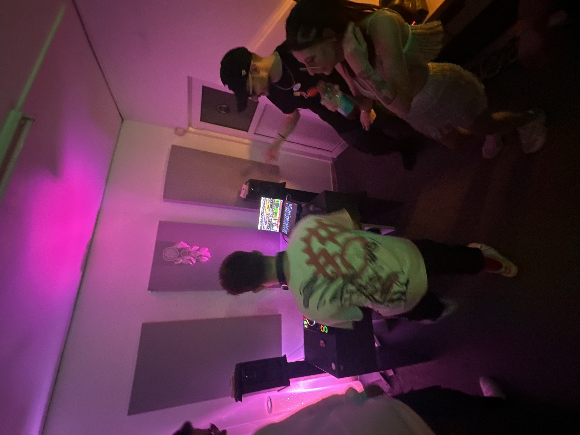 An indoor scene depicts four individuals within what appears to be a music studio or production room, characterized by dominant pink and purple ambient lighting emanating from overhead. The room features white walls, a dark floor, and grey rectangular acoustic panels mounted on the left and far walls. A white door with a dark handle and a small window is visible on the right wall.
Central to the scene, a male subject with dark, curly hair, wearing a light-colored t-shirt with a complex graphic design on the back and headphones around his neck, stands facing a black DJ mixer/controller. The mixer displays illuminated green and orange buttons. To his immediate left, a black studio monitor is positioned on a stand. Behind the mixer, a laptop computer sits on a stand, displaying a colorful digital interface.
To the right of the central subject, a male subject wearing a black baseball cap, dark sunglasses, and a black t-shirt with visible graphics (depicting "The Simpsons" characters) gestures with his right hand. Adjacent to him, a female subject with light brown hair and glasses holds a blue bottle with a straw in her right hand, while her left arm is wrapped around the male subject's lower back. Both are looking towards the left side of the frame.
Partially visible on the far left, a fourth male subject, wearing a light-colored top and dark pants, observes the scene. Wall decor includes a dreamcatcher-style object with feathers, positioned between two acoustic panels. No explicit visual cues within the image identify its specific location as being within the city of Plovdiv, Bulgaria.