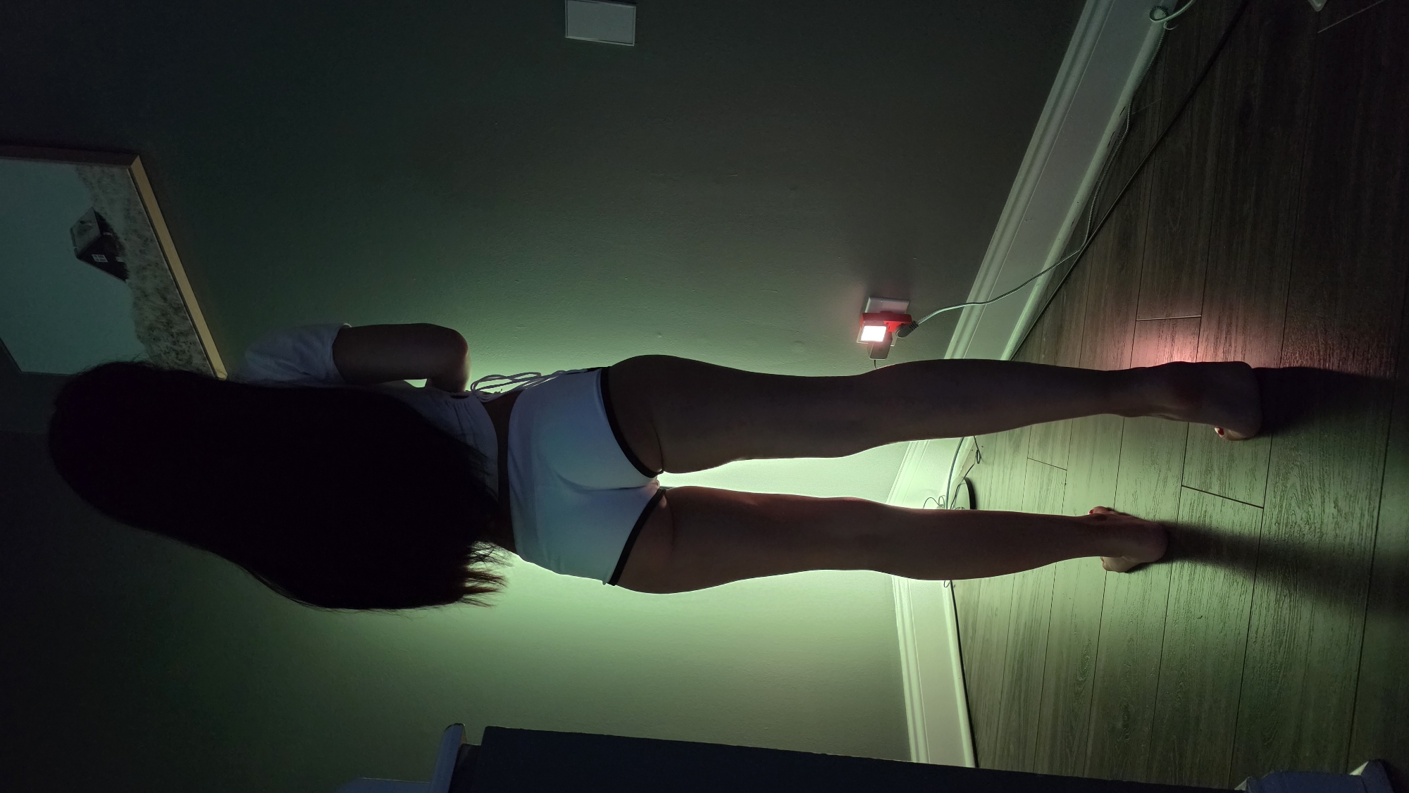 A person is standing in a room with dark wood floors and a green-lit wall. The person is facing away from the camera, with their back to the viewer. They have long, dark hair and are wearing a white crop top and white shorts with black trim. The lighting in the room is dim, with a green glow emanating from the wall behind the person, and a small red light is visible on the wall near the floor. A framed picture of a house on a hill is hanging on the wall. The overall impression is of a dimly lit, indoor setting. There is no information in the image to suggest a location in the city of Elgin, United States of America.