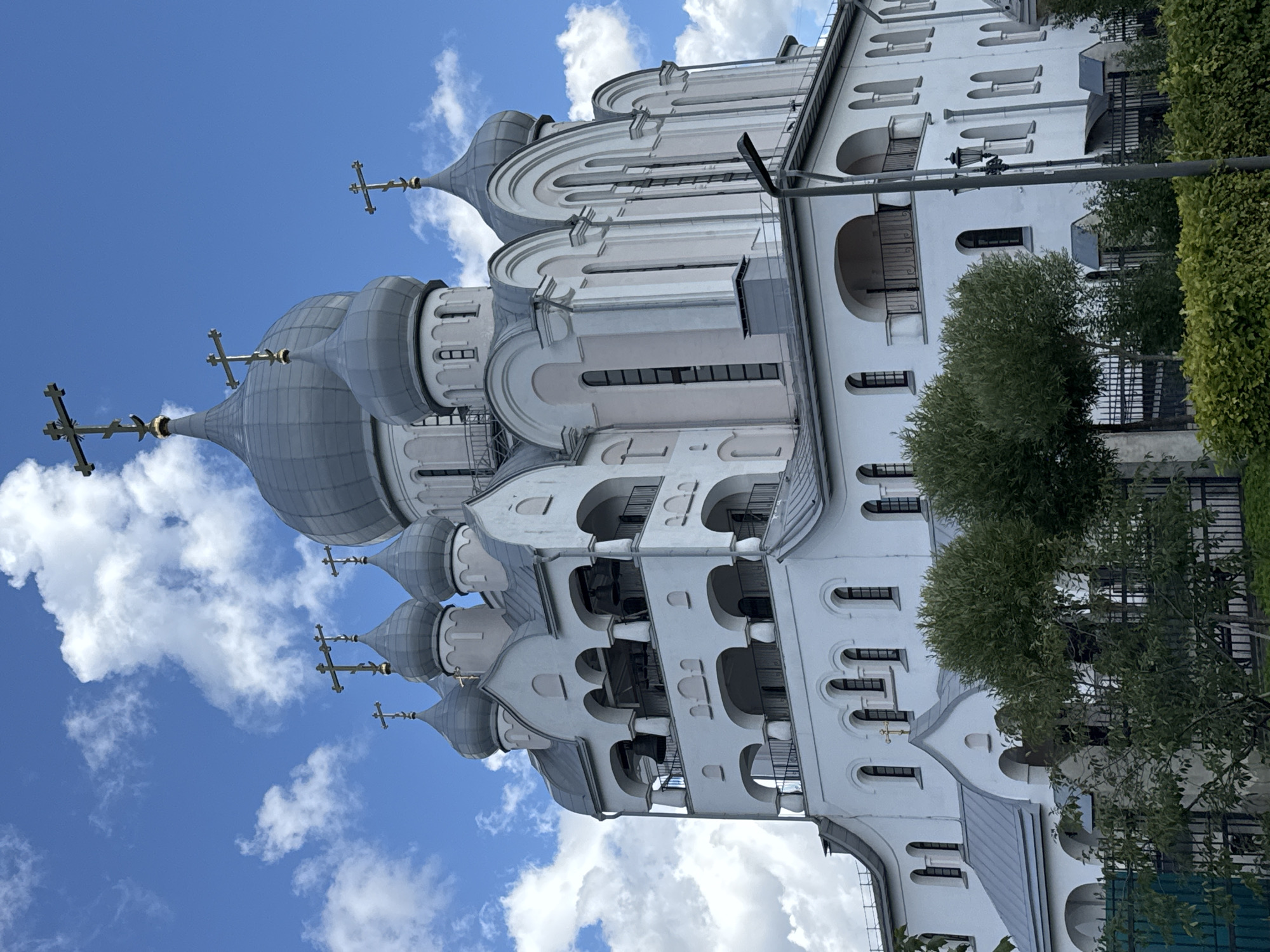 The image shows a large, white Orthodox church with multiple domes topped with crosses, set against a blue sky with scattered white clouds. The church has an elaborate design with arched windows and balconies, and a bell tower. There are trees and fences in the foreground. The church is located in Saint Petersburg, Russia. The specific location is the Church of the Resurrection of Christ, also known as the Savior on Spilled Blood.
