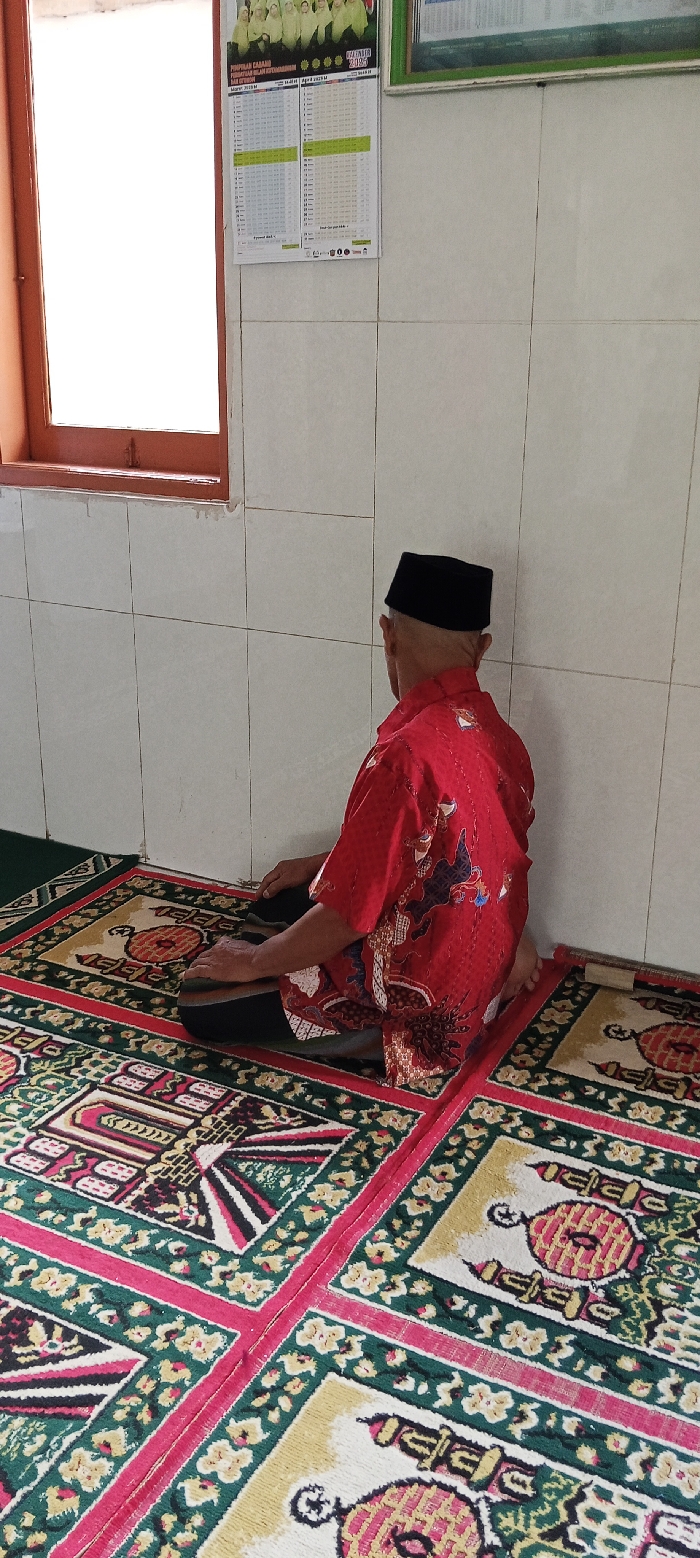 A man wearing a red and black patterned shirt and black hat is kneeling on a prayer rug. He is facing a white tiled wall with a window in the background. The window is covered by a brown frame and is mostly obscured by sunlight. A calendar hangs on the wall. There is another prayer rug partially visible in the foreground. The scene is set inside a room in a building located in Kecamatan Kutawaringin, Indonesia.