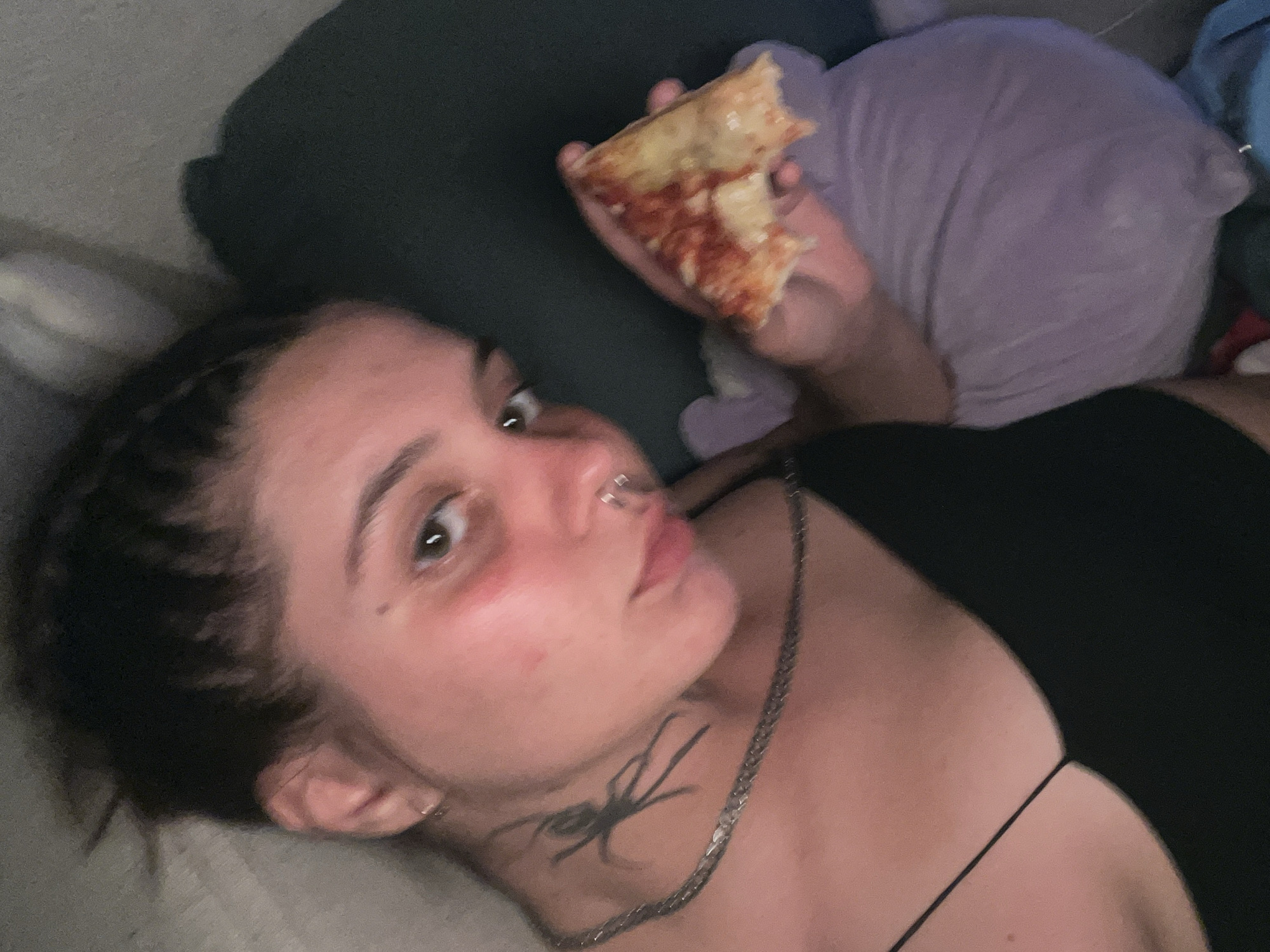 A young woman with dark hair styled in braids, wearing a black spaghetti strap top and a silver chain necklace with a spider pendant, lies down with a slice of pizza in her right hand. She has a nose ring and a spider tattoo on her neck. Her left cheek appears flushed. She is holding the pizza over a purple stuffed toy. The background is dimly lit, with a hint of a gray wall or furniture. The image is a close-up, selfie-style shot taken indoors. There is no information available to determine the location in the city of Genemuiden, Netherlands.