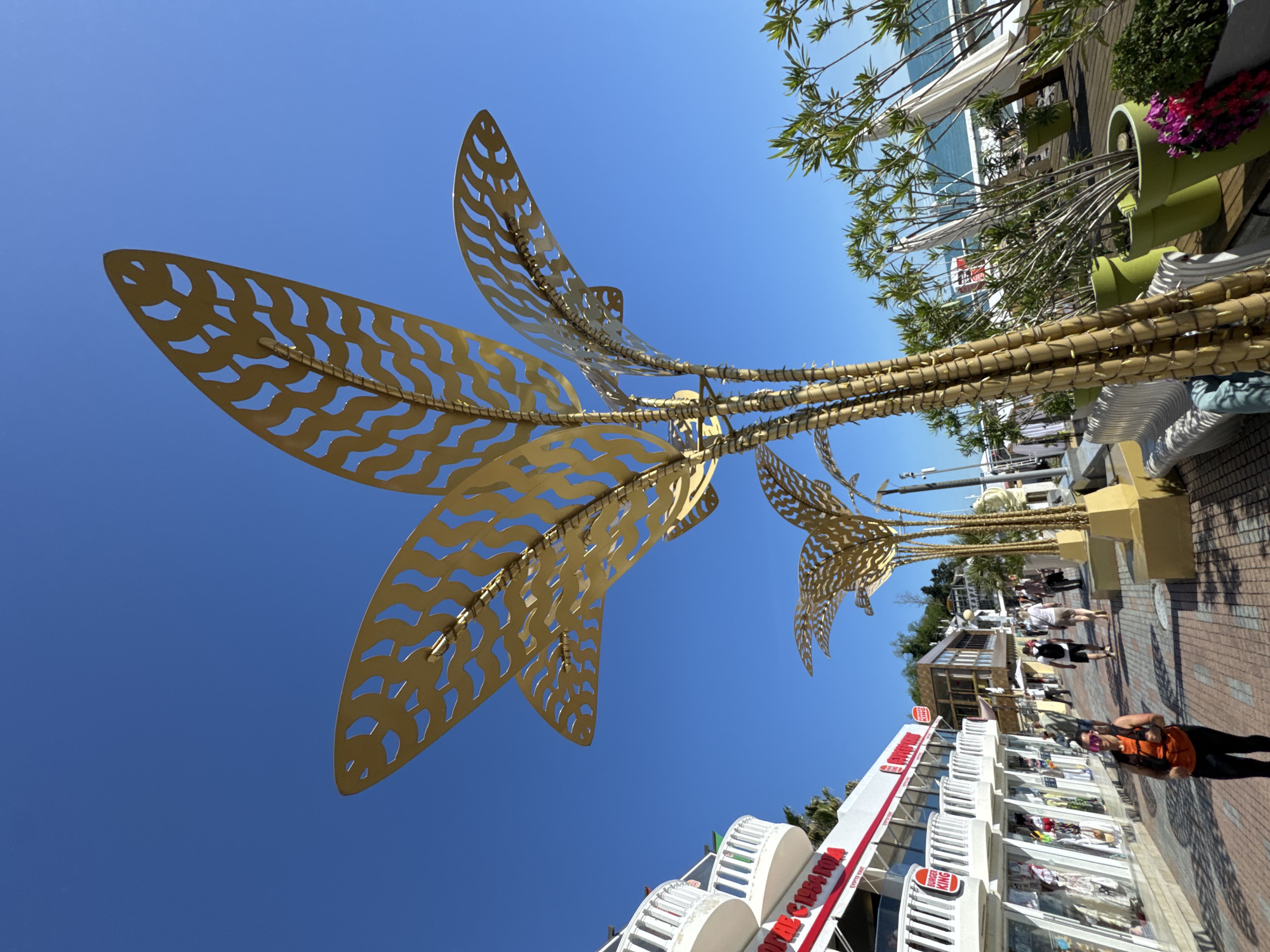 This image captures a vibrant, sunny scene along a commercial promenade in Sochi, Russia. The most prominent subjects are a series of large, golden metallic sculptures resembling stylized palm trees or tropical foliage, positioned against a clear blue sky. These sculptures feature intricate, wave-like cut-out patterns on their broad fronds and are supported by multi-branched, golden-colored structures that mimic tree trunks.
Below the sculptures, a bustling pedestrian street is visible, lined with multi-story buildings housing shops and establishments. The buildings are predominantly white with red accents, featuring large display windows and some balconies. Several people are seen walking along the brick-paved street, dressed in casual summer clothing. In the foreground, a person wearing an orange top and black pants walks past the shops.
Potted plants in large green containers and real trees with green foliage add greenery to the scene, some of which appear to be olive trees or similar species. The bright sunlight indicates it is daytime, likely mid-day or early afternoon, with perfectly clear skies.
Visible text on the buildings includes "BURGER KING" on multiple signs and "ГАСТРОНОМЪ" (Gastronom) in red lettering on a larger sign, with "EVERGREEN" written below it. The overall atmosphere suggests a lively tourist or shopping area under pleasant weather conditions.