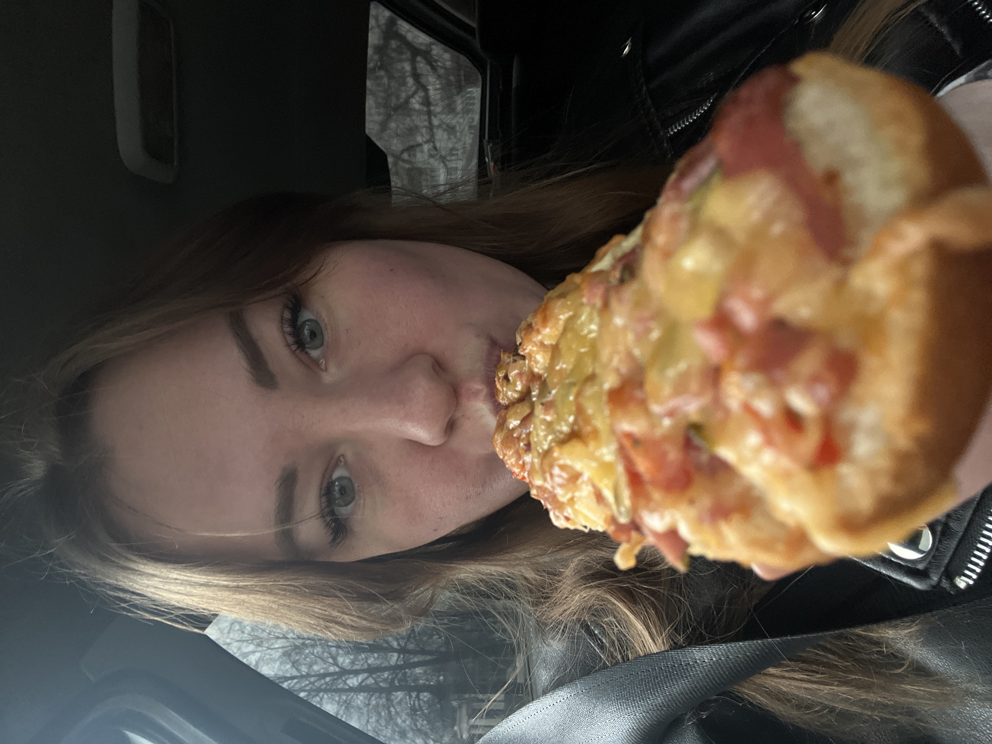 A young woman with light brown hair and fair skin is eating a loaded bread roll or pizza-like snack in what appears to be a car. She is holding the food up to her mouth with her right hand, suggesting she is about to take a bite. The snack is topped with melted cheese and what seems to be meat, such as sausage or ham. The woman is wearing a black leather jacket. The background shows the interior of a car and a blurry view through the window, possibly trees. The location cannot be determined without additional information or context.