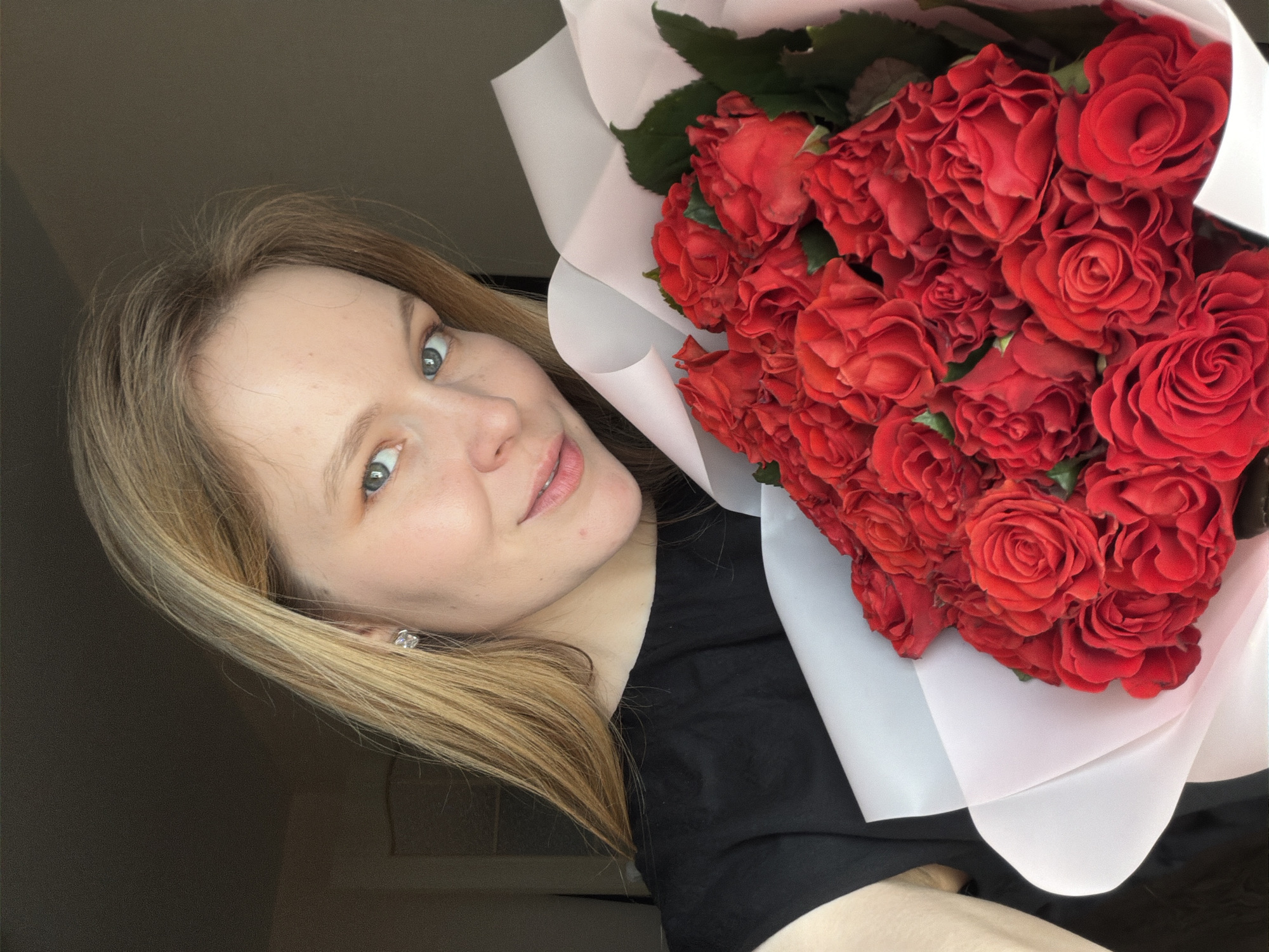 A light-skinned woman with shoulder-length blonde hair, wearing a black top and a small, square earring, is holding a large bouquet of red roses wrapped in white paper. The roses are densely packed, with visible green leaves among the blossoms. The background is out of focus, but suggests an indoor environment with a gray wall. The image is rotated slightly counter-clockwise. Location: Izhevsk, Russia not determined.
