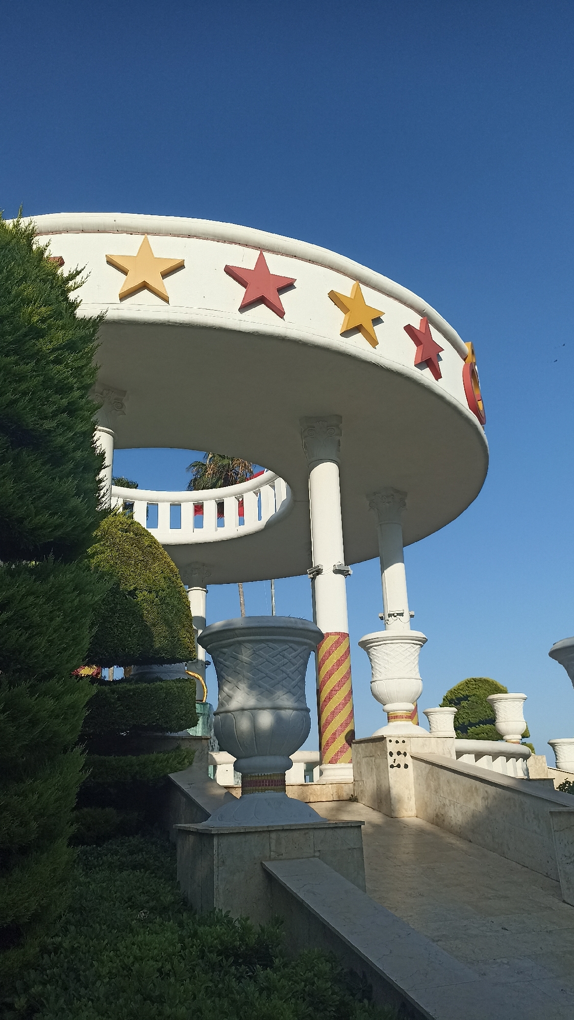 The image captures a bright, sunny day in Mezitli, Turkey, showcasing an ornate, circular structure against a clear blue sky. The structure, likely a pavilion or gazebo, features a white, curved roof adorned with alternating yellow and red stars. It is supported by white columns. In the foreground, lush green bushes and trees flank the entrance to the structure, which appears to be part of a landscaped public space. Large, decorative white planters with intricate patterns are strategically placed along a tiered, marble-like walkway leading up to the pavilion. The walkway and planters suggest a well-maintained and decorative outdoor area, possibly a park or a recreational facility within Mezitli. The overall scene conveys a sense of vibrancy and public leisure.