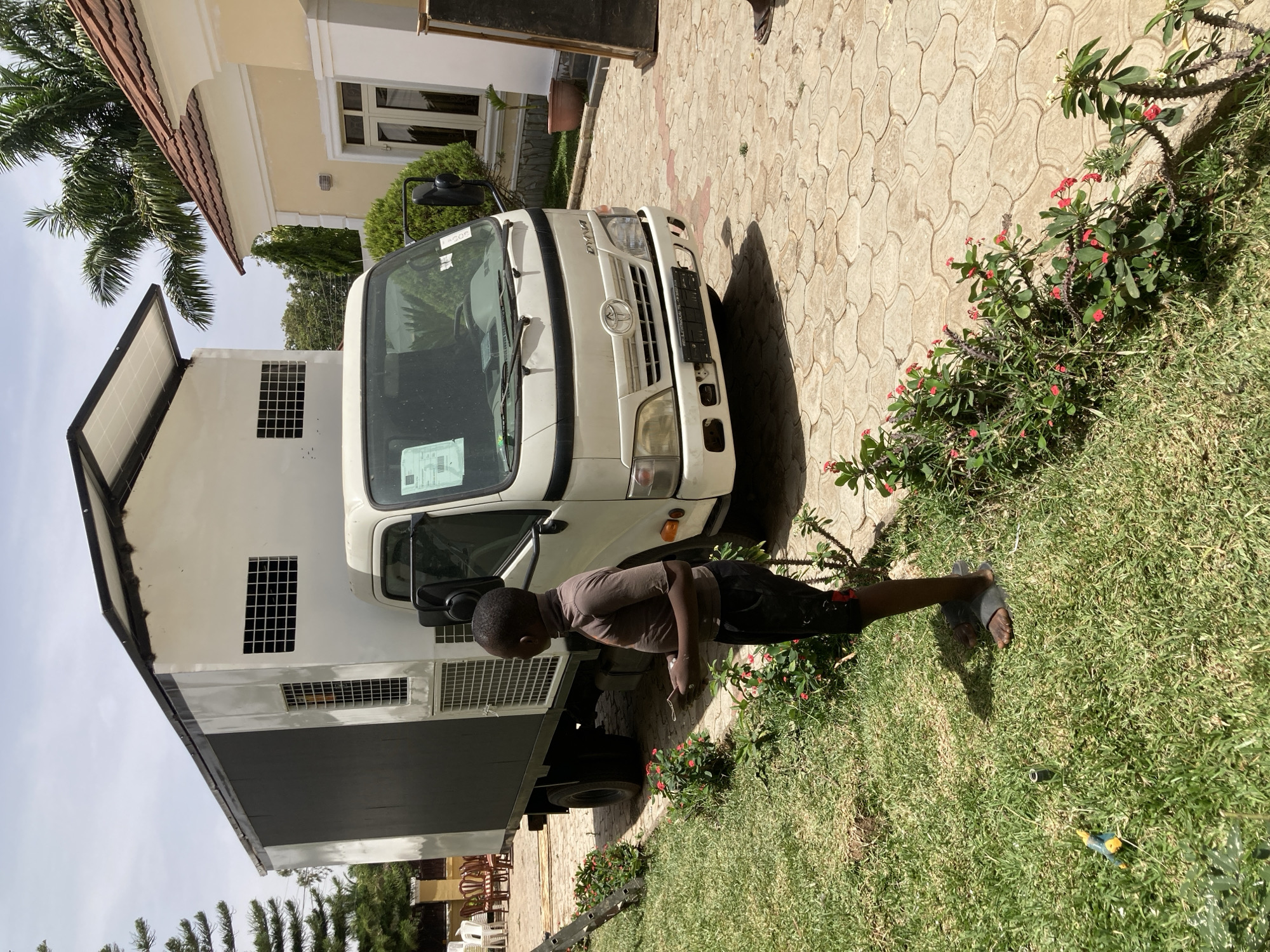 The image depicts a scene in Jos, Nigeria. A light-skinned Black boy wearing a brown shirt and black shorts stands on a patch of green grass beside a white truck with a black cargo container. The truck is parked on a patterned concrete driveway. The boy appears to be holding something in his hands. The driveway is flanked by green foliage with small red flowers. Behind the truck, a two-story white building with a dark roof is visible. A portion of a beige building with a red-tiled roof is also visible in the background, along with a palm tree. In the background, there are dark green trees. The weather appears to be sunny and bright.