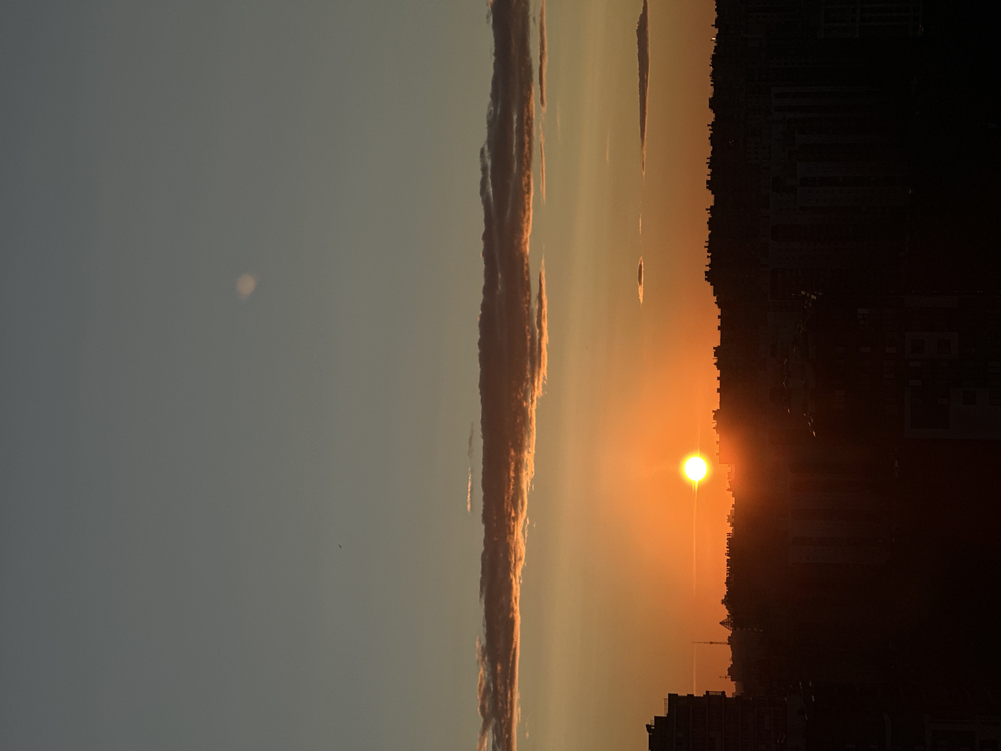 The image captures a sunset over a cityscape. The sky transitions from a pale blue at the top to a warm orange hue closer to the horizon. A thin, elongated cloud band with illuminated edges stretches horizontally across the middle of the frame. The sun, a bright orange orb, is partially visible, positioned slightly to the left of the center, casting a warm glow and creating lens flare effects. Below the sky, a dark silhouette of a city skyline is visible, with the tops of buildings and structures forming an irregular line. A tall, dark building is prominent on the left edge of the frame. The lower portion of the image is dominated by the darkened silhouettes of urban buildings. The scene is devoid of any people or specific actions. The overall impression is of a tranquil evening in an urban environment.