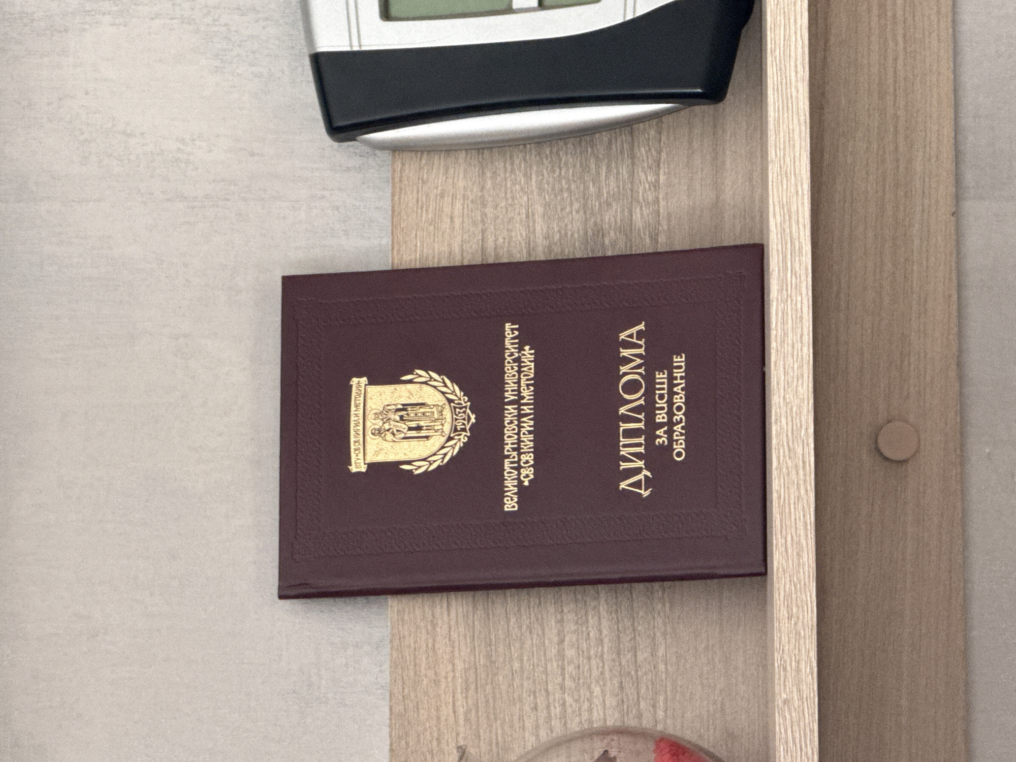 The image shows a diploma from Veliko Tarnovo University "St. St. Cyril and Methodius" in Bulgaria. The diploma is maroon with gold lettering and is partially obscured in a shelf with another object above it. The diploma has the university's seal and the year 1967 on it. It says "DIPLOMA" and "FOR HIGHER EDUCATION" in Cyrillic. The shelf is made of light-colored wood. The location cannot be determined from the image.