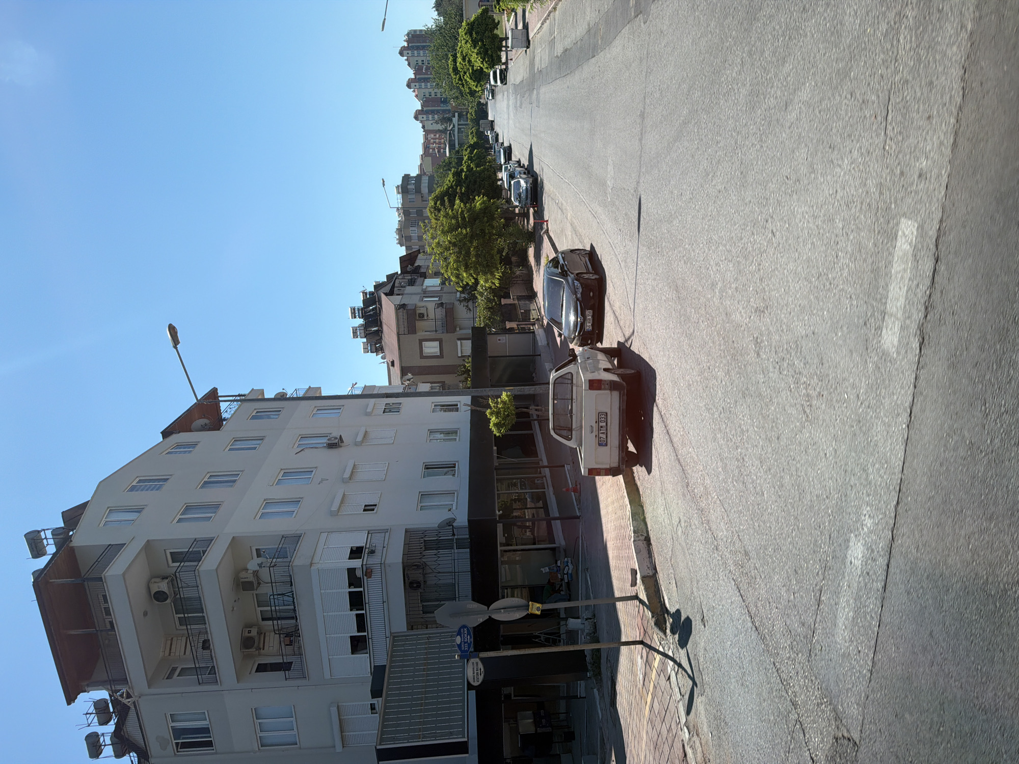 The image captures a street scene in Konyaaltı, Turkey, on a clear, sunny day. The perspective is from street level, looking down a moderately trafficked road lined with multi-story buildings and trees.
In the foreground, a white pickup truck with a license plate reading "07 ATM 033" is parked on the right side of the road. To its left, a black sedan with license plate "46 AAR 950" is also parked. The road surface is asphalt, showing some signs of wear and tear with visible seams and patches. A sidewalk made of interlocking pavers is visible on the left, bordered by a curb. A street sign with text including "333. SOKAK" is present near the sidewalk.
The buildings lining the street are modern, with several floors, balconies, and air conditioning units visible. Some buildings have solar water heaters on their roofs. Trees with green foliage are interspersed between the buildings, providing shade. In the background, more buildings and a clearer view of the sky are visible. The overall environment suggests a residential or mixed-use urban area. No people are clearly visible in the image. The lighting indicates it is daytime, likely late morning or early afternoon, given the strong shadows cast.