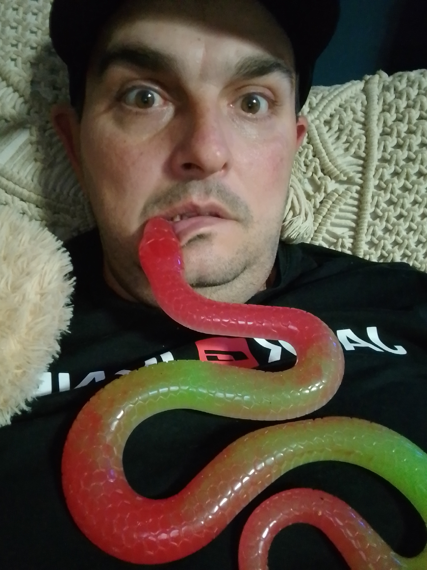 A man wearing a black baseball cap and a black t-shirt with white lettering on it, is lying down, looking at the camera. A red and green gummy snake is draped across his chest, with its head in his mouth. The man has wide eyes and his tongue is sticking out slightly. A light-colored fuzzy object is visible on the left side of the image, and a textured blanket is behind the man. The man appears to be indoors, but no specific location in Wedel, Germany is discernible from the image.