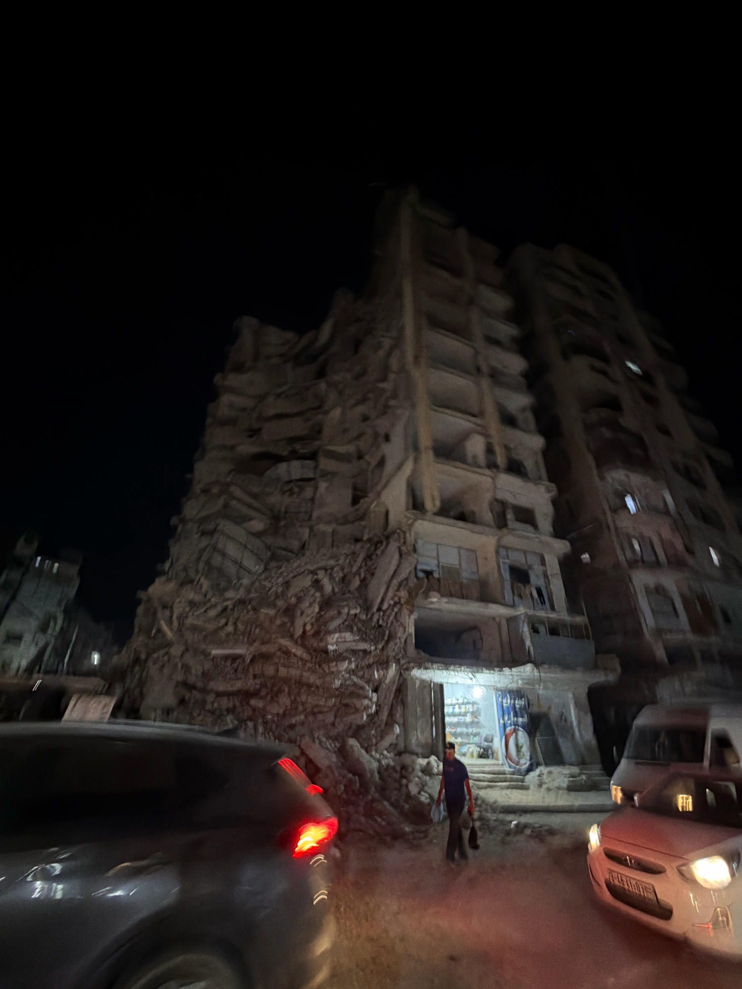 The image captures a nighttime scene in Gaza City, Palestinian Territories, showcasing the aftermath of destruction. In the foreground, the rear of a dark-colored vehicle is visible, its red taillights illuminated. To the right, a white car with its headlights on is also present.
Dominating the midground and background is a severely damaged multi-story building. The left side of the building has collapsed, with large sections of debris and rubble piled up at its base. The remaining structure appears precarious, with exposed floors, damaged walls, and broken windows. The building's facade is weathered and shows signs of significant structural compromise.
A person, dressed in dark clothing with a blue shirt, is walking in the debris-strewn area in front of the damaged building. This individual is carrying bags, suggesting they may be scavenging or transporting goods. Behind them, a dimly lit storefront is visible, with shelves stocked with items. The overall environment is dark, with artificial lighting from the vehicles and the storefront casting a reddish glow on the surrounding rubble. The scene conveys a sense of devastation and the ongoing challenges faced in the aftermath of conflict.
