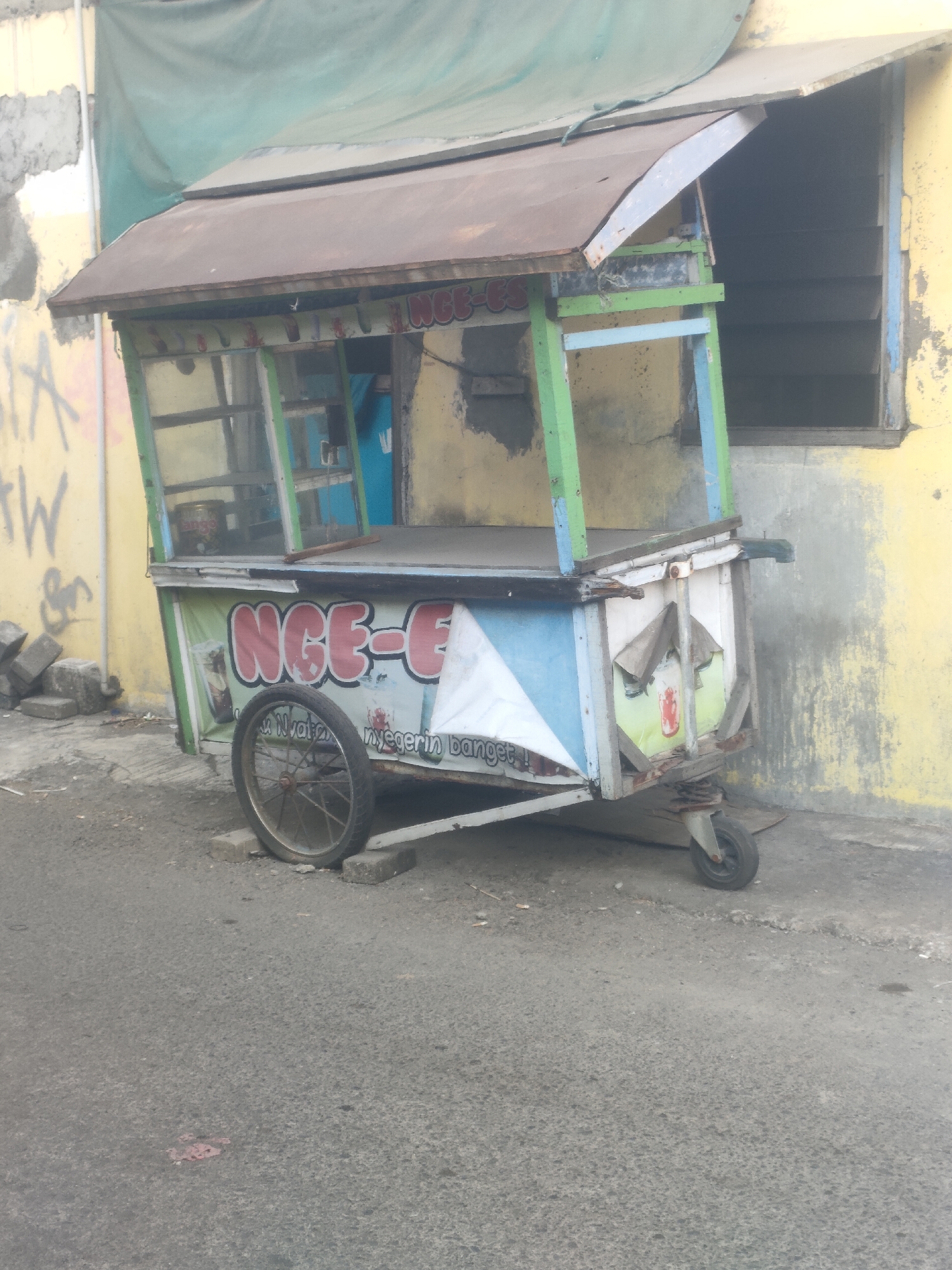 The image shows an abandoned food cart with a rustic design, likely in Surabaya, Indonesia. The cart is made of wood and metal, with a tilted roof covered by a blue tarp. It has a single large wheel at the front and a smaller wheel at the back, suggesting it was once mobile. The cart's facade is decorated with colorful graphics, including the text "NGE-E" and an image of a drink. The cart is positioned on a paved street, with a yellow-painted building in the background, marked with graffiti. The scene appears to be in a state of disrepair, with some debris scattered around the cart. The lighting suggests it is daytime, possibly overcast.