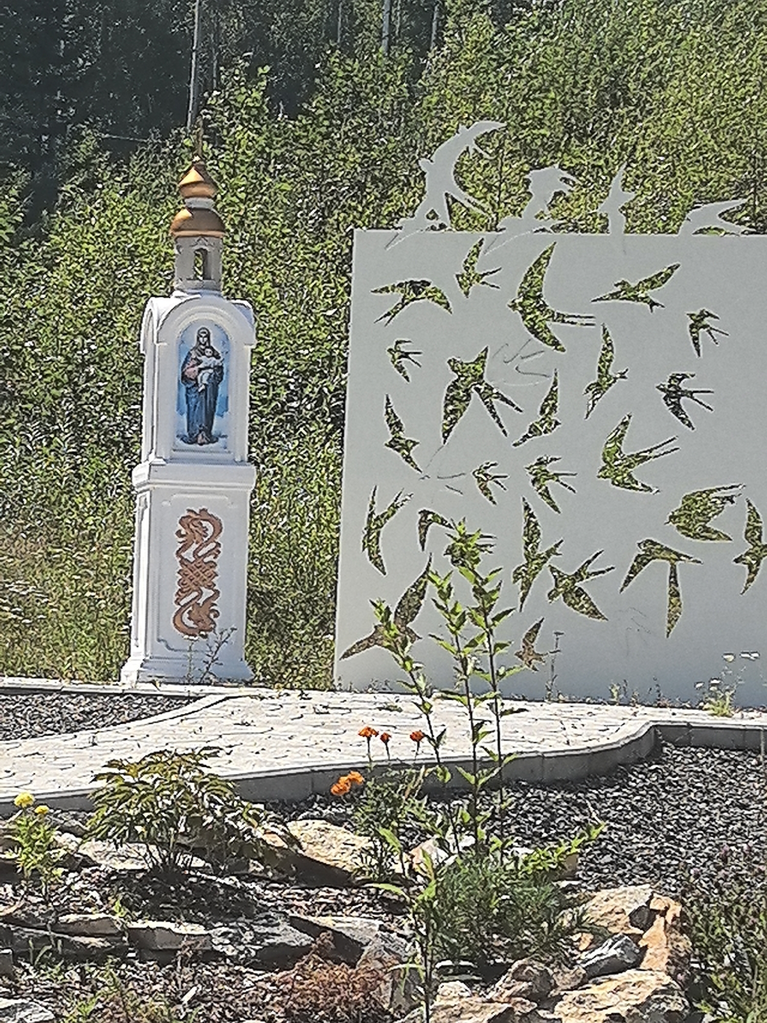 The image shows a memorial or wayside shrine featuring a statue of the Virgin Mary holding the infant Jesus, situated next to a large, white panel decorated with cut-out silhouettes of birds in flight. The shrine is white and rectangular, with a gold-colored domed roof and decorative brown scrollwork on its base. The white panel, also rectangular, has a pattern of numerous bird silhouettes, some of which are filled with a green, textured material, possibly to resemble foliage or camouflage. The scene is set outdoors, with lush green foliage and trees in the background. In the foreground, there is a paved pathway made of light-colored stones, leading towards the shrine and the panel. The ground around the pathway is covered with dark gravel and scattered rocks, with some low-lying plants and flowers. The overall impression is of a tranquil outdoor space, possibly a garden or a memorial park. The specific location in the city of Solnechnyi, Russia is not visually discernible from the image itself.