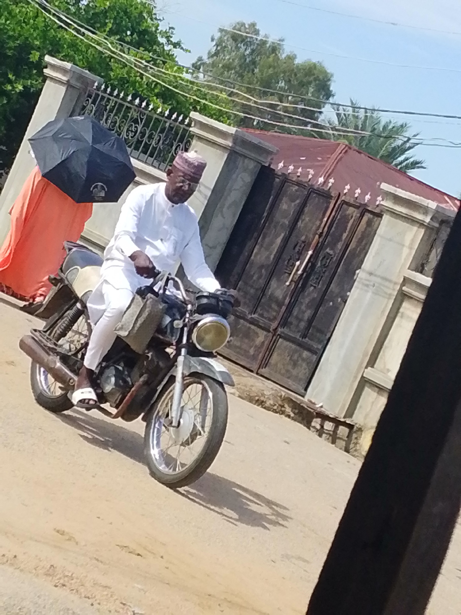 A man wearing a white shirt and pants, sandals, and a small cap is riding a motorcycle on a paved road. He is holding onto the handlebars, appearing to steer the motorcycle. Beside him to the left, there's a person in an orange gown holding a black umbrella. Behind them, a building has a large gate and a brown roof. The scene appears to be on a street, likely in Lokoja, Nigeria, given the architectural style and clothing. The setting seems to be a sunny day.