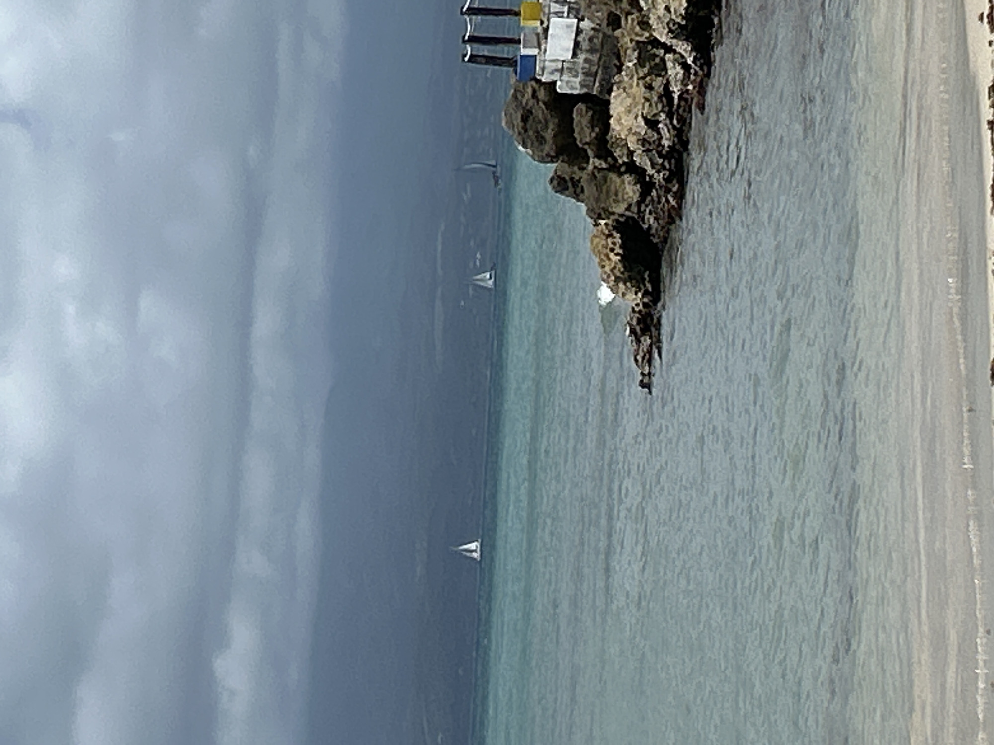 The image shows a coastal scene under a cloudy sky. The location is Unknown, Unknown. There's a rocky outcrop with a structure on top, possibly a small building or marker, painted with white, blue, and yellow sections. The sea is a light turquoise color, with at least three sailboats visible in the distance. The right side of the image shows a beach with light sand.