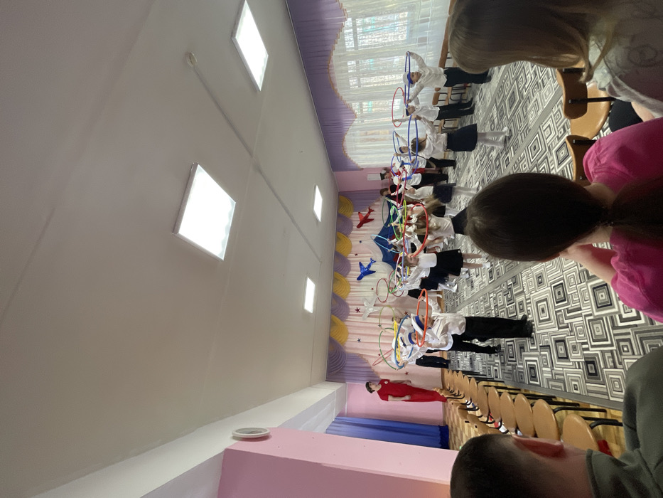 Children in matching white shirts and dark skirts or pants stand in rows on a patterned floor, holding colorful hoops. A woman in a red dress stands near chairs lined up for an audience. A stage backdrop features airplane decorations. The room has high ceilings with square lights. Viewers' heads are visible in the foreground.