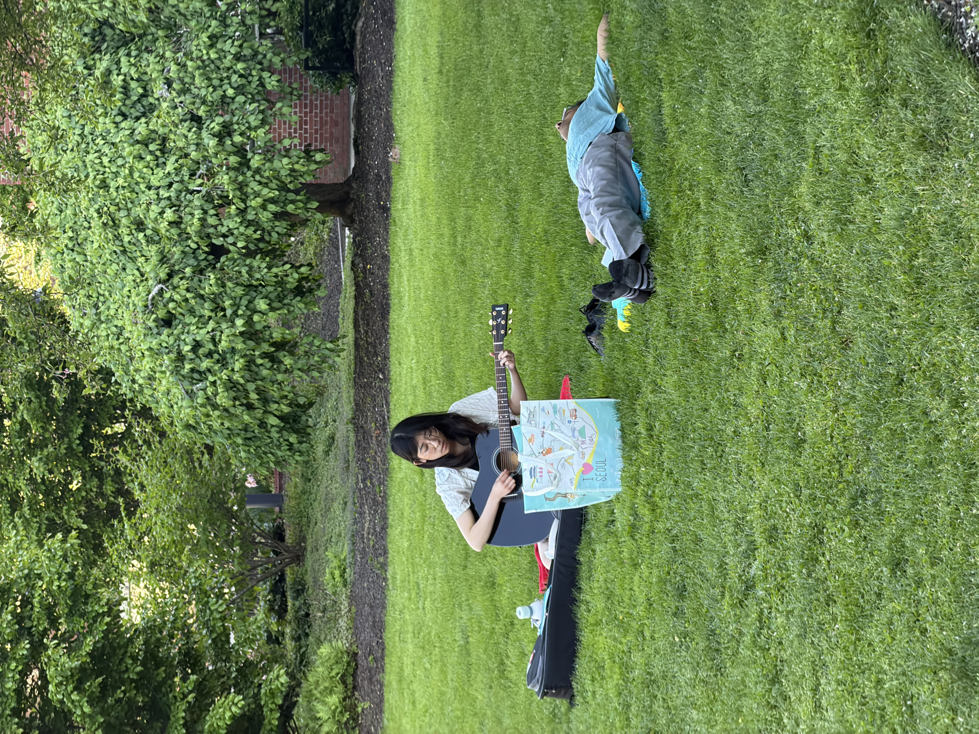 A woman sits on the grass in what appears to be a park or yard, holding a guitar and a shopping bag that reads "I <3 SEOUL." Next to her is a guitar case and a water bottle. A young person lies in the grass nearby with one arm raised. A tree stands to the left of the woman, and a brick building can be seen in the background. Given the possible clues in the description of the location, it is possible to assume this image was taken in Cambridge, Massachusetts, United States of America.