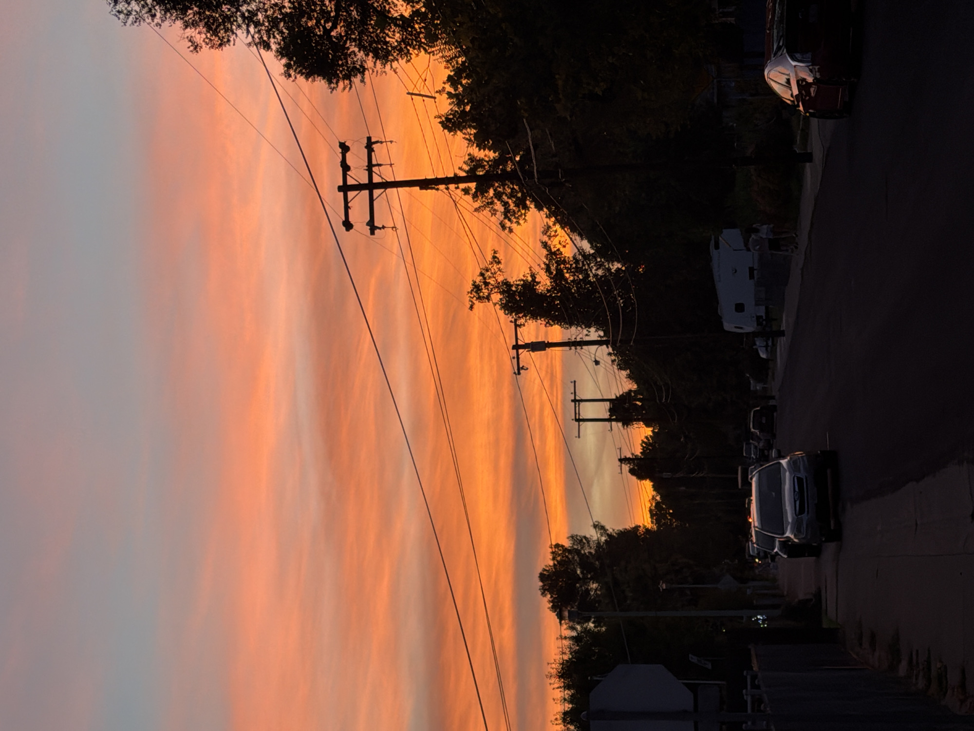 The image depicts a street scene at sunset in Sacramento, California. The sky is filled with vibrant hues of orange and pink, with wispy clouds stretching across the horizon. Telephone poles with numerous wires are silhouetted against the colorful sky, casting long shadows on the street.
Several cars are parked along the side of the road and driving down the street. In the foreground, a silver SUV is the most prominent vehicle. Further down the street, a red sedan is visible on the right side of the road.
On the left side of the street, a fence runs along the sidewalk, separating the road from a property. Trees line the street on both sides, creating a sense of depth and framing the scene. In the distance, a white RV or camper is parked. Two people are standing near the RV, appearing to interact with each other.
The overall atmosphere of the image is tranquil and serene, capturing the beauty of a sunset in an urban environment. The composition draws the viewer's eye towards the colorful sky and the silhouetted telephone poles, while also capturing the details of the street and the vehicles.